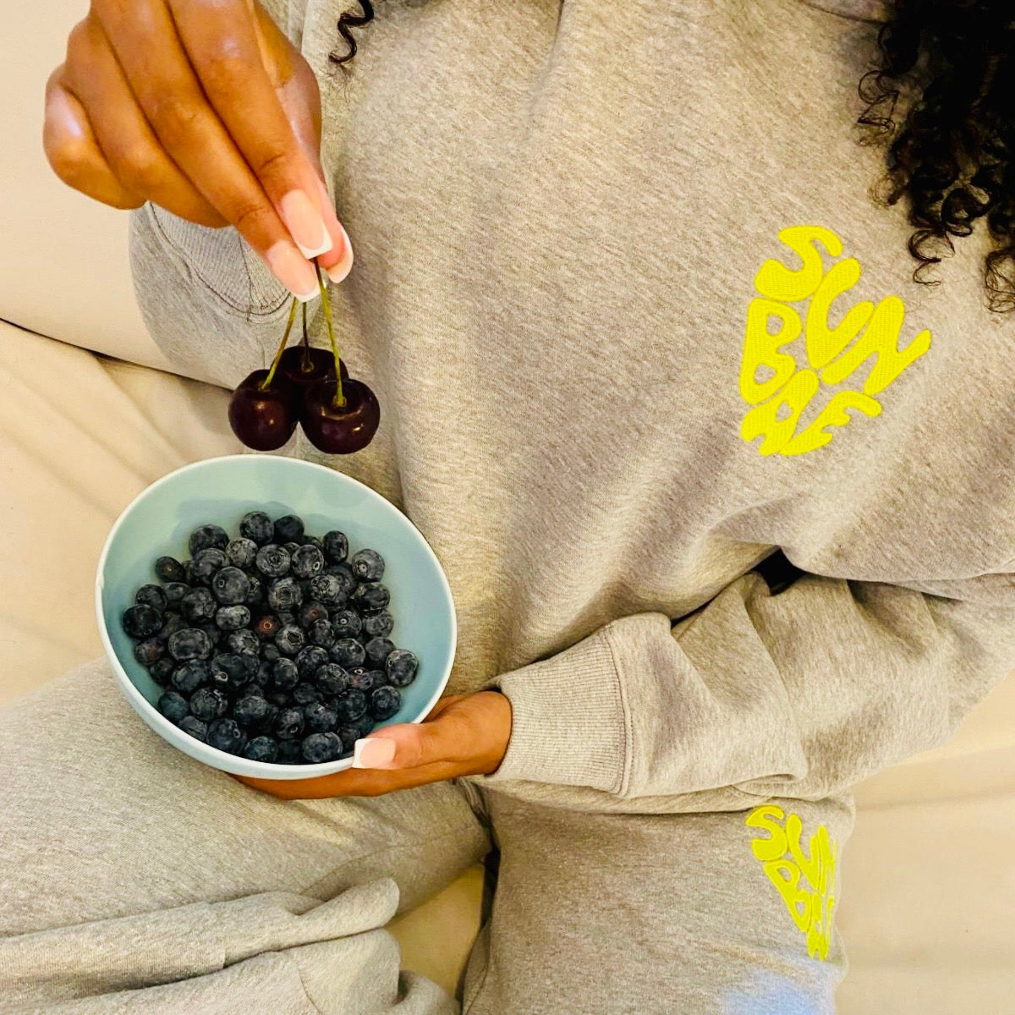 Person holding a bowl of blueberries and cherries, wearing a grey SUNBAE Hoodie and Sweat Pant.
