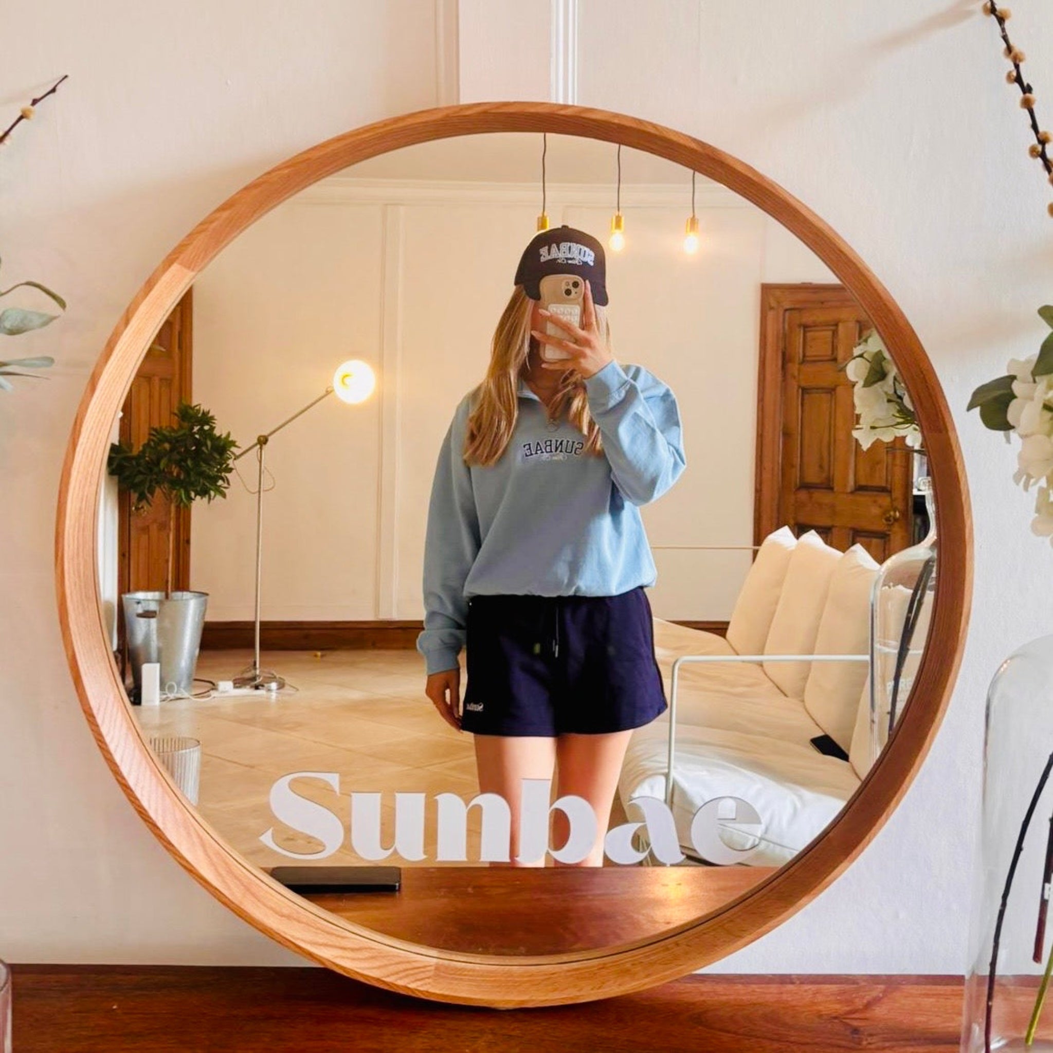 Sunbae Glower wearing SUNBAE Cloudburst Zip Sweat with Varsity Embroidery in Blue taking a mirror selfie in a room with a round wooden-framed mirror.