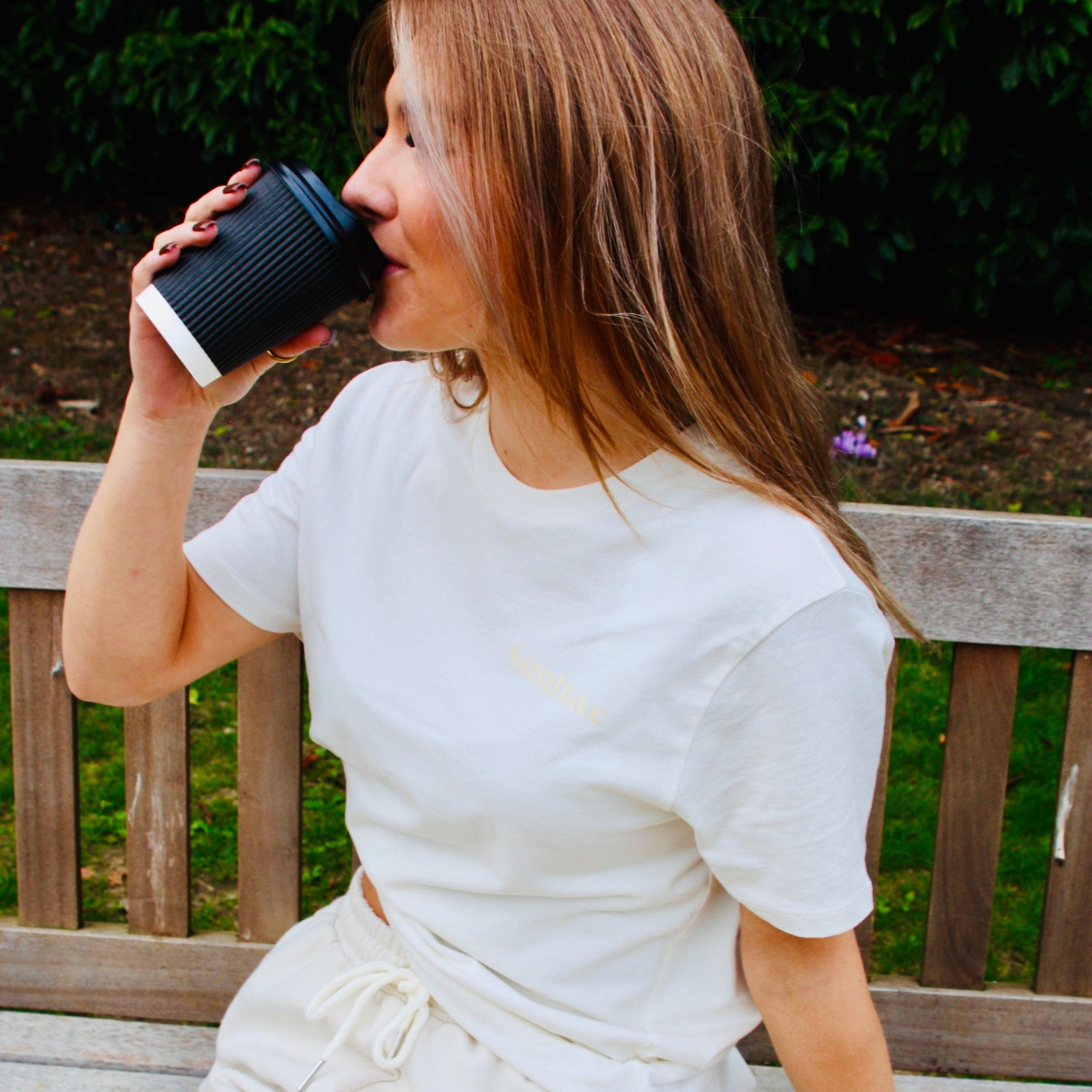 Woman drinking from a black coffee cup while sitting on a wooden bench outdoors.