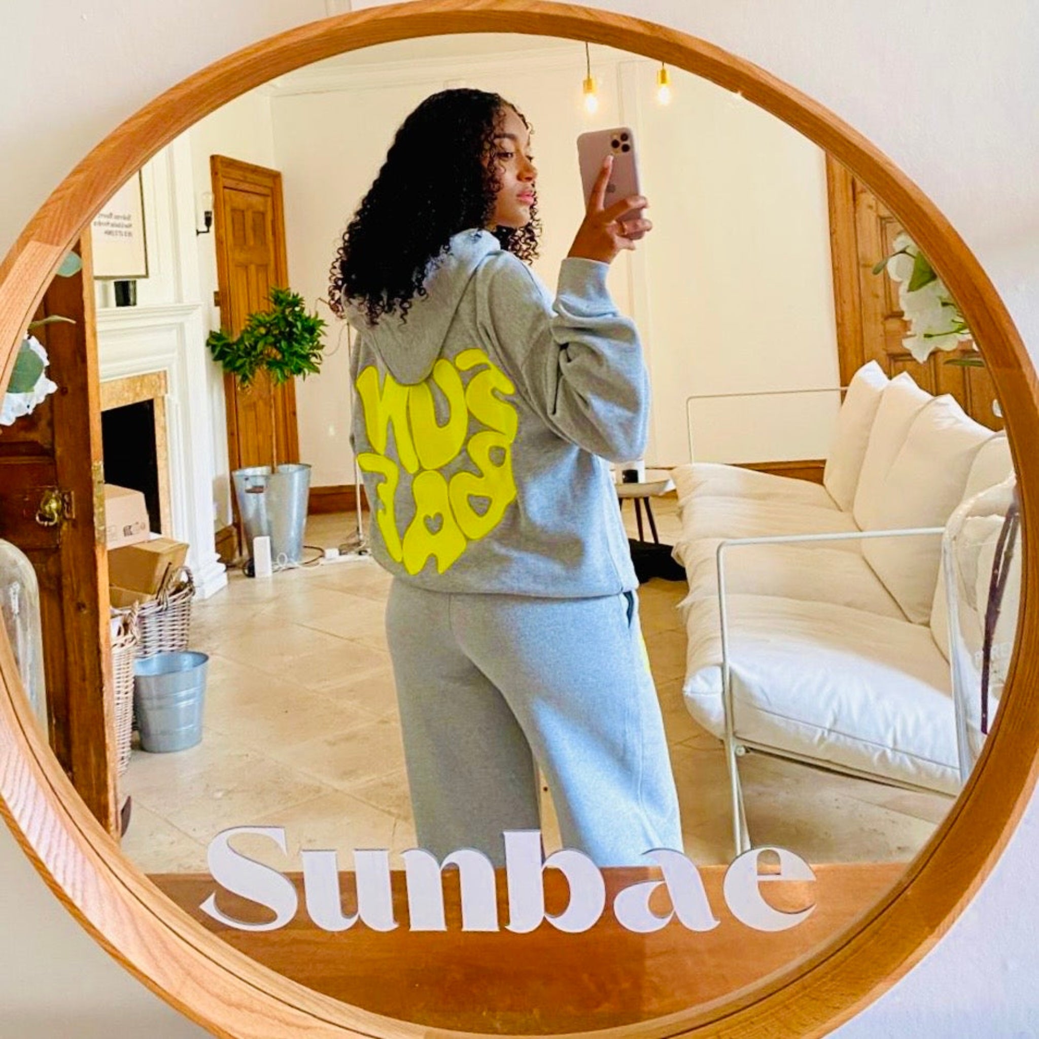 Person taking a mirror selfie wearing a SUNBAE grey hoodie in a living room.