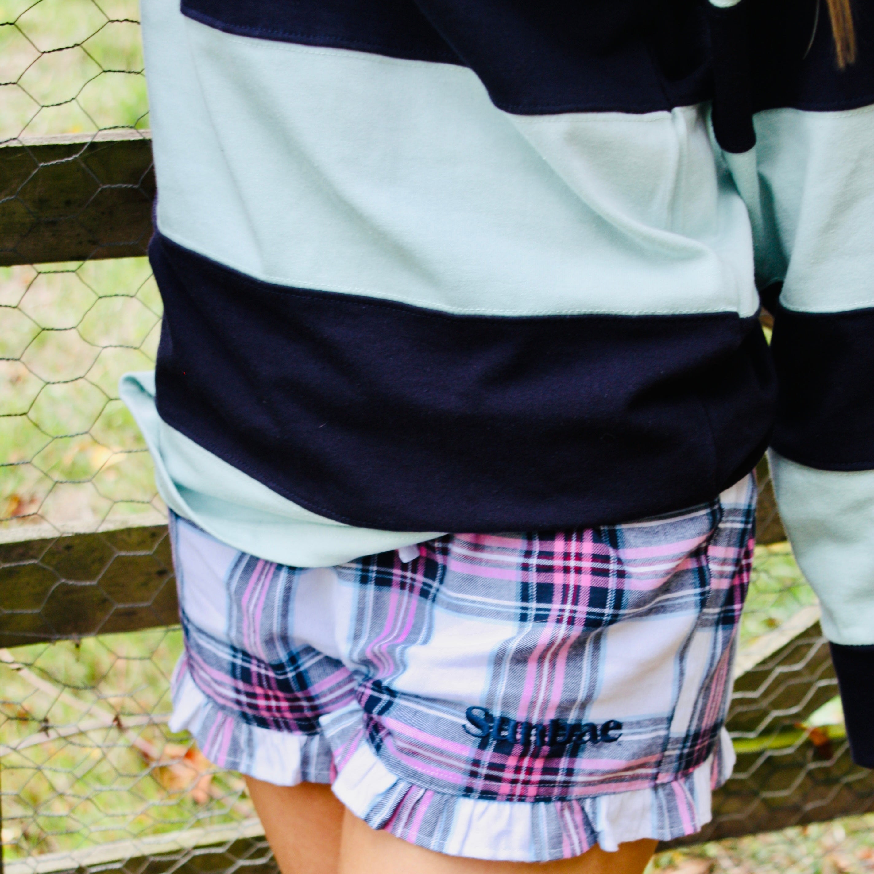 Person wearing a plaid skirt with 'Sunshine' text in front of a metal fence.