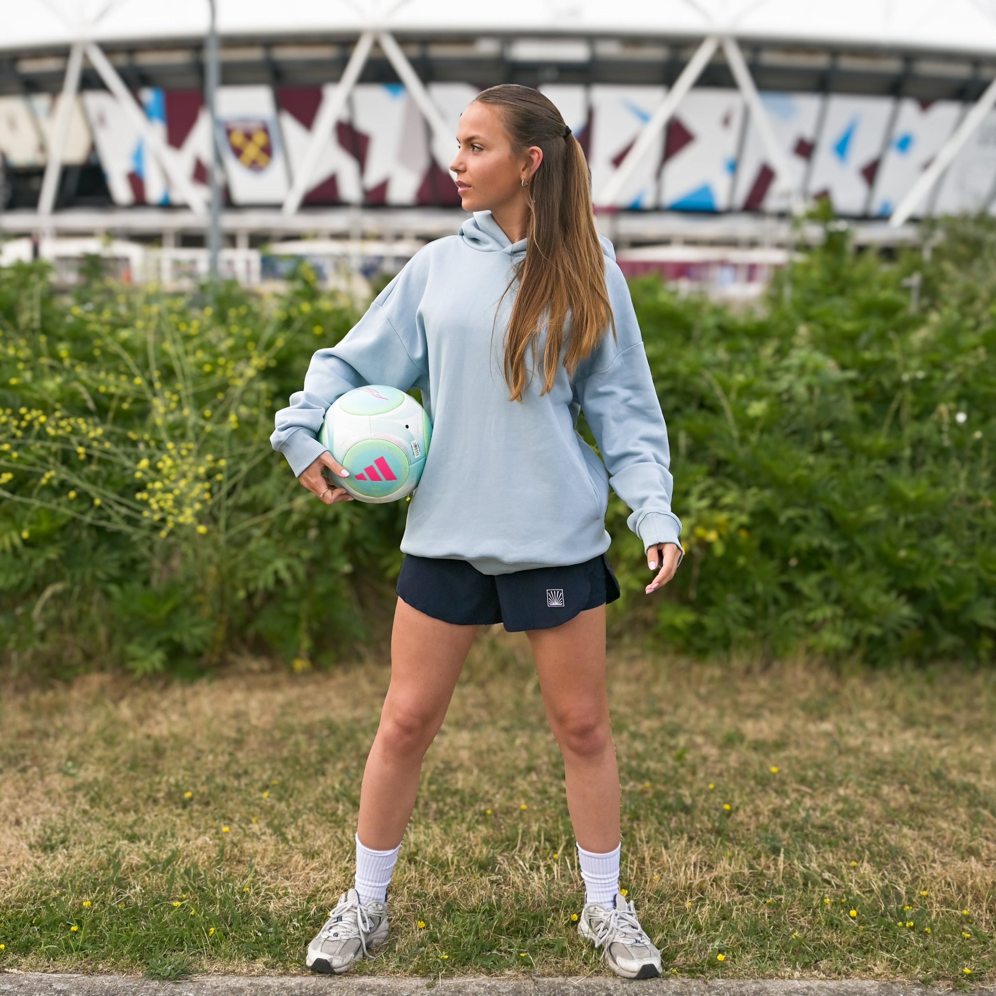 Yvonne Main wearing a Sunbae hoodie and holding a soccer ball in front of a stadium