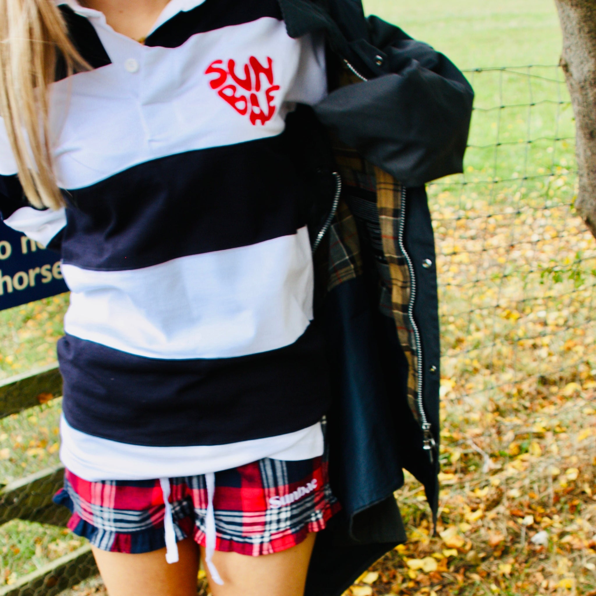 Person wearing a striped shirt, plaid skirt, and rain boots in an outdoor setting with leaves on the ground.