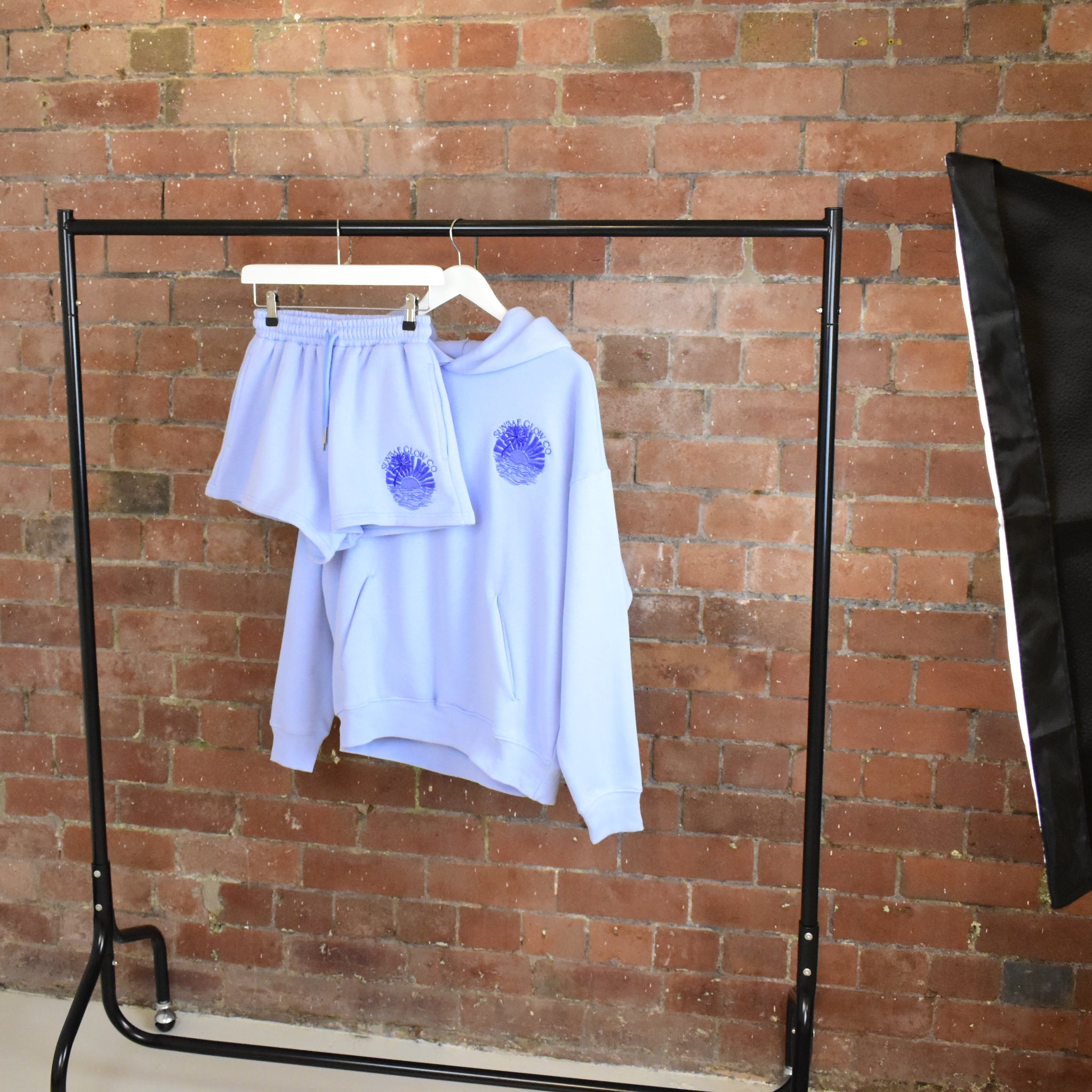 Light blue SUNBAE Hoodie and Short set on a black rack against a brick wall.