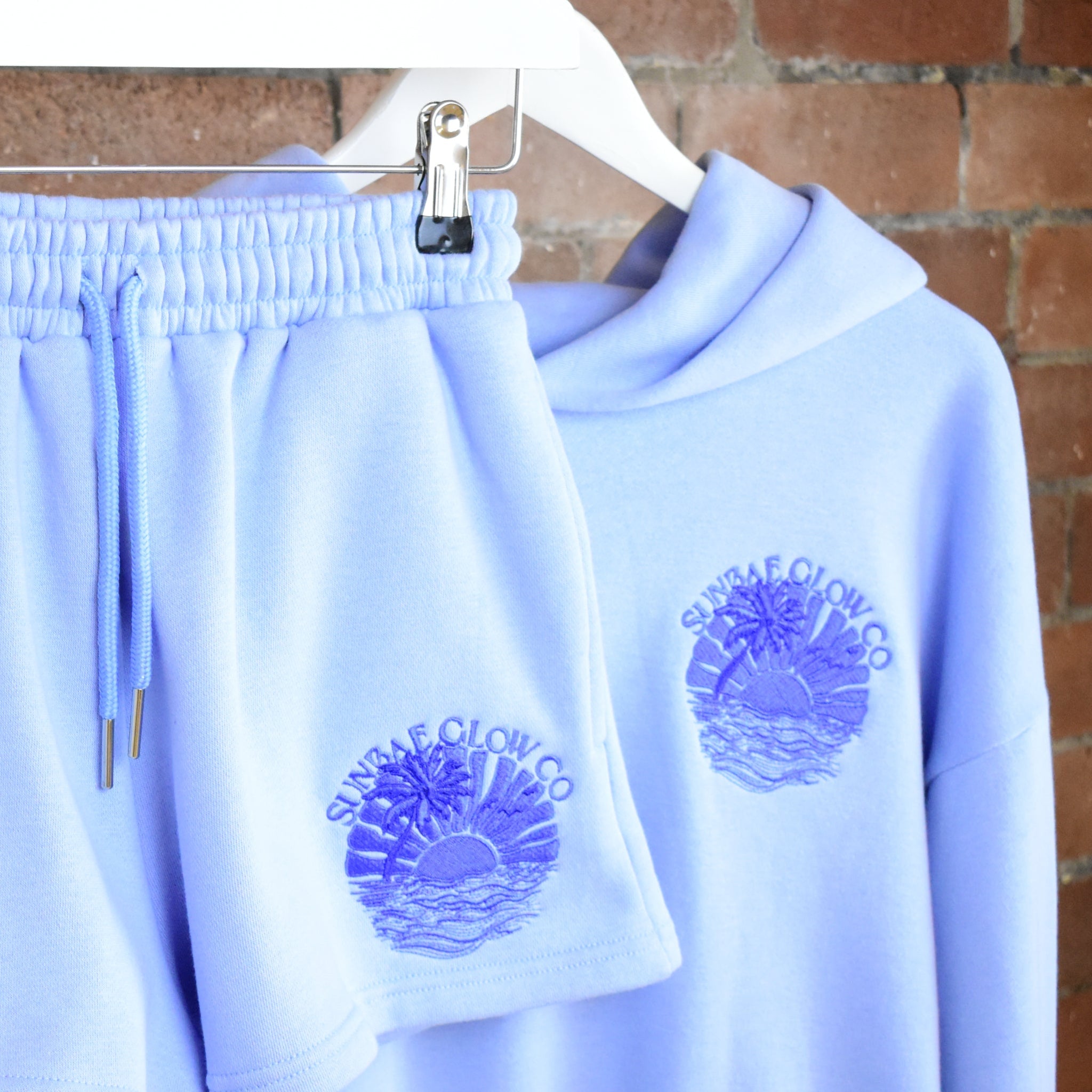 SUNBAE Tidal Glow Hoodie and Short Co-ord set with 'Sunset Glow Co.' logo on a hanger against a brick wall.