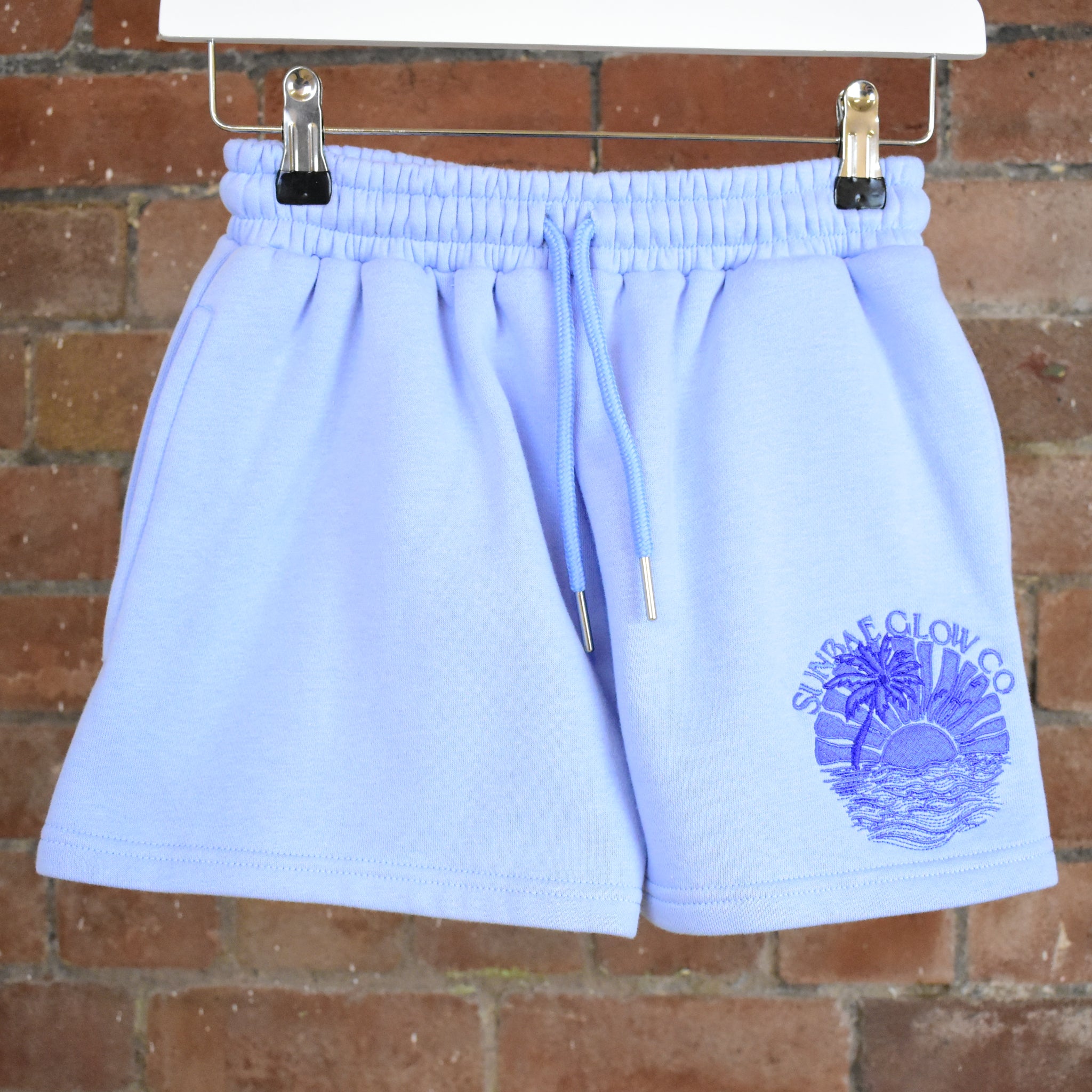 SUNBAE Light blue shorts with a logo on a brick wall background