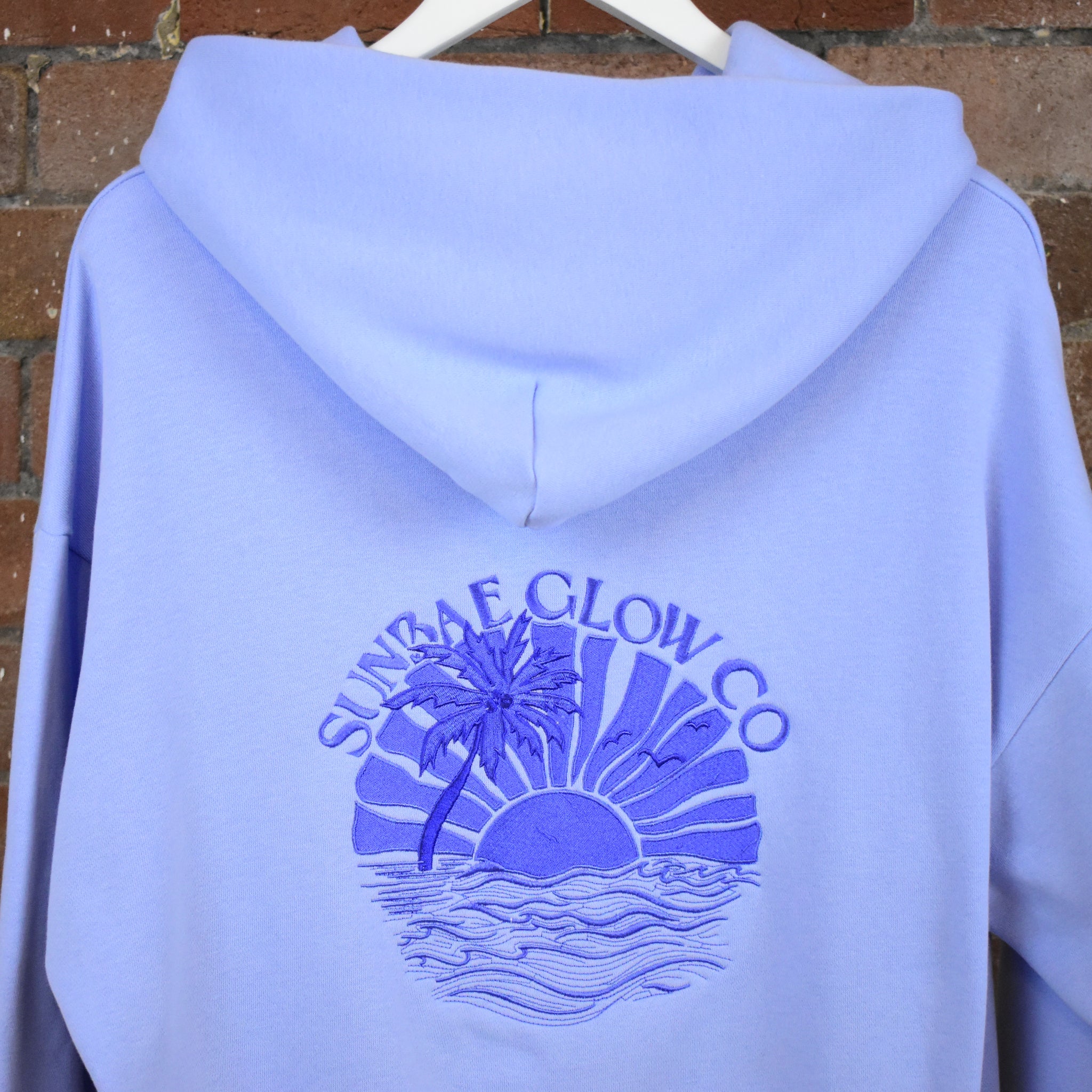 Back of a SUNBAE Tidal Glow hoodie with a palm tree and sun design on a brick wall background