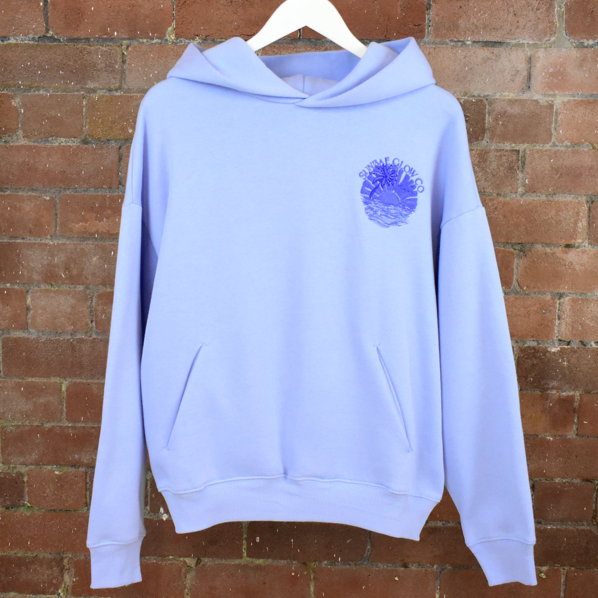 SUNBAE Tidal Glow hoodie with a logo on a hanger against a brick wall