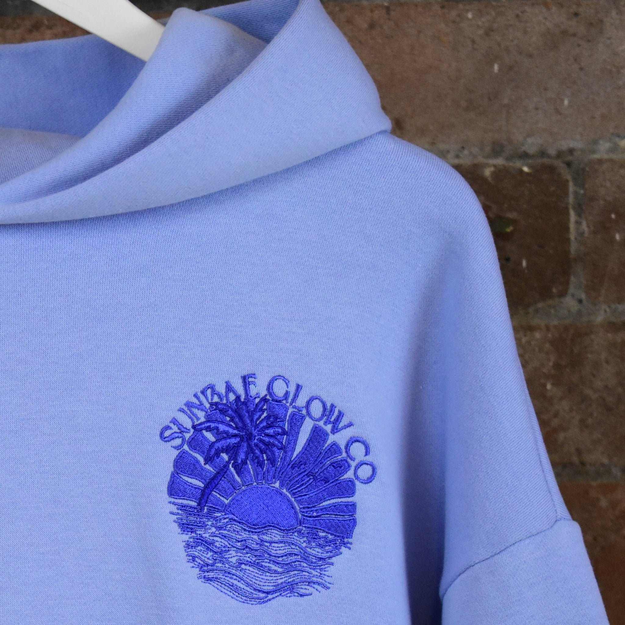 Blue SUNBAE hoodie with embroidered design against a brick wall