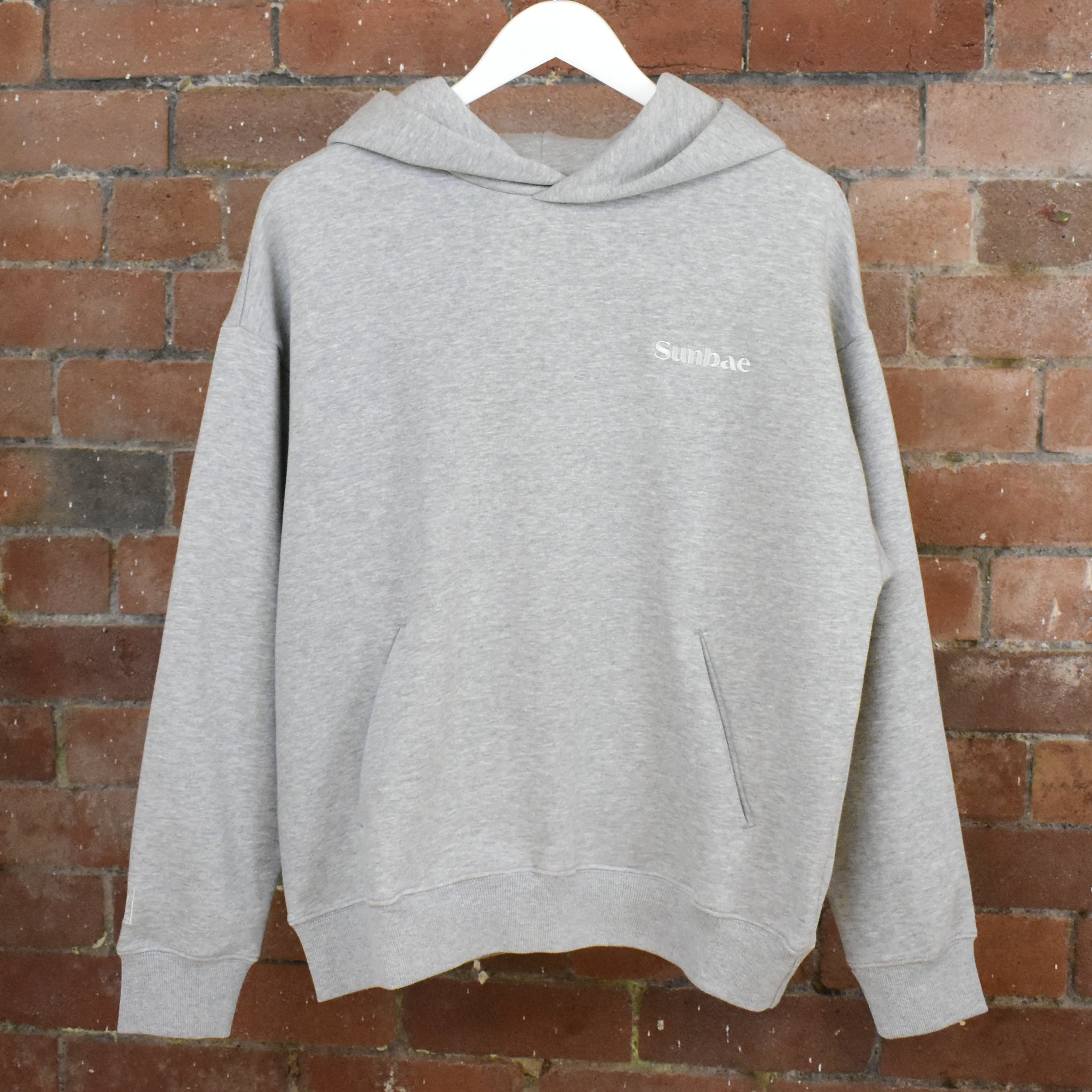 Grey Sunbae hoodie with a brand logo on a hanger against a brick wall
