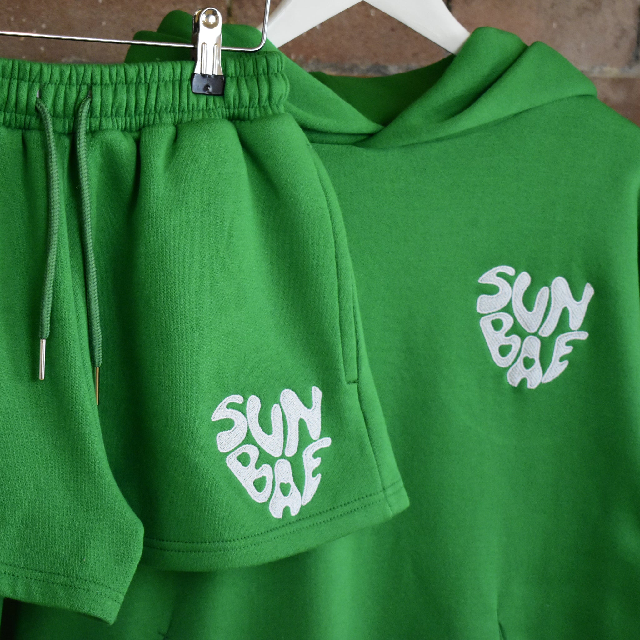 Green hoodie and shorts set with SUNBAE logo on a hanger against a brick wall.