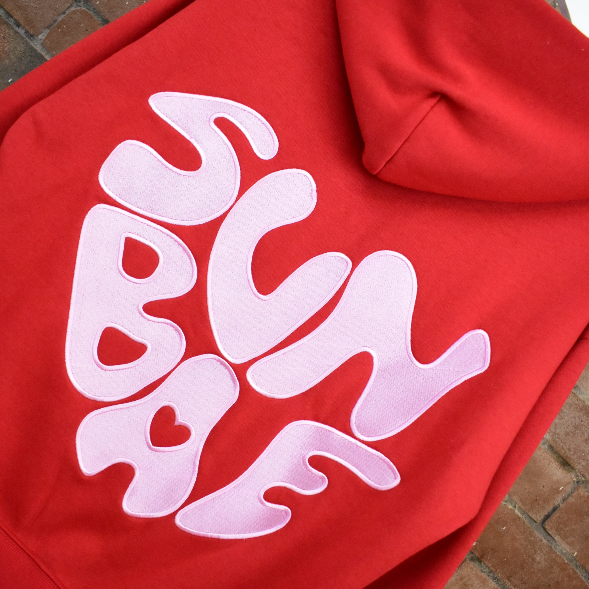 Red hoodie with pink SUNBAE logo on a brick surface