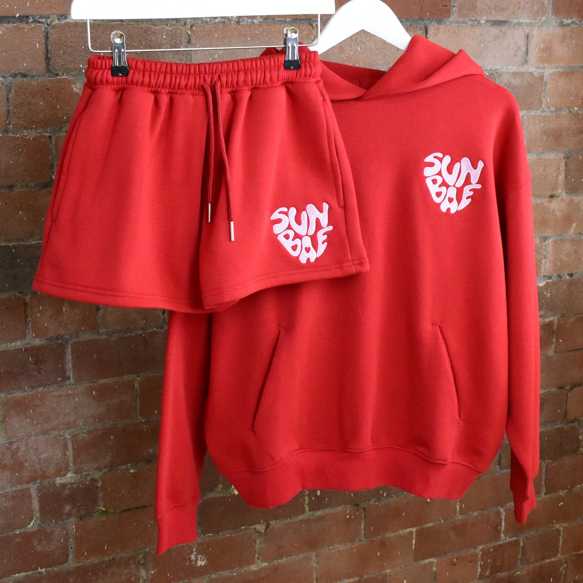 Red SUNBAE Glazed Heart hoodie and shorts set on a brick wall background