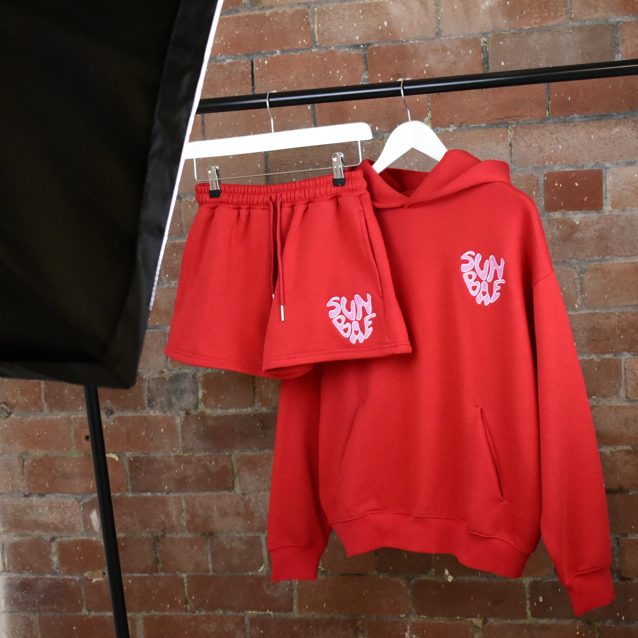 Red SUNBAE hoodie and shorts set with on hangers against a brick wall.