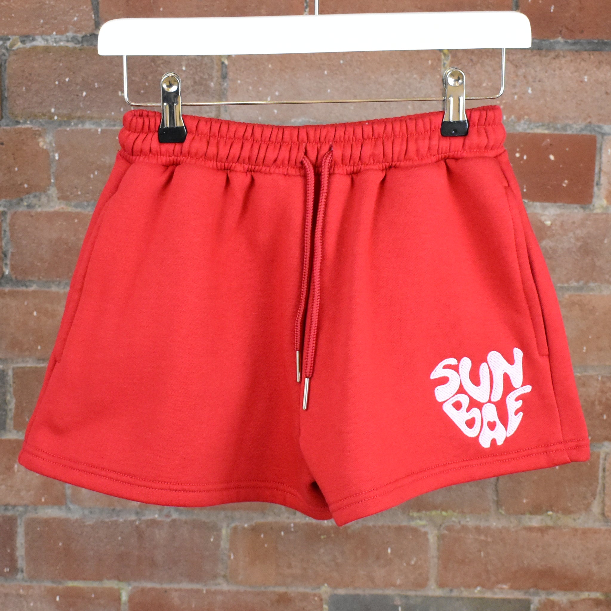 Red SUNBAE Glazed Heart shorts on a hanger against a brick wall.