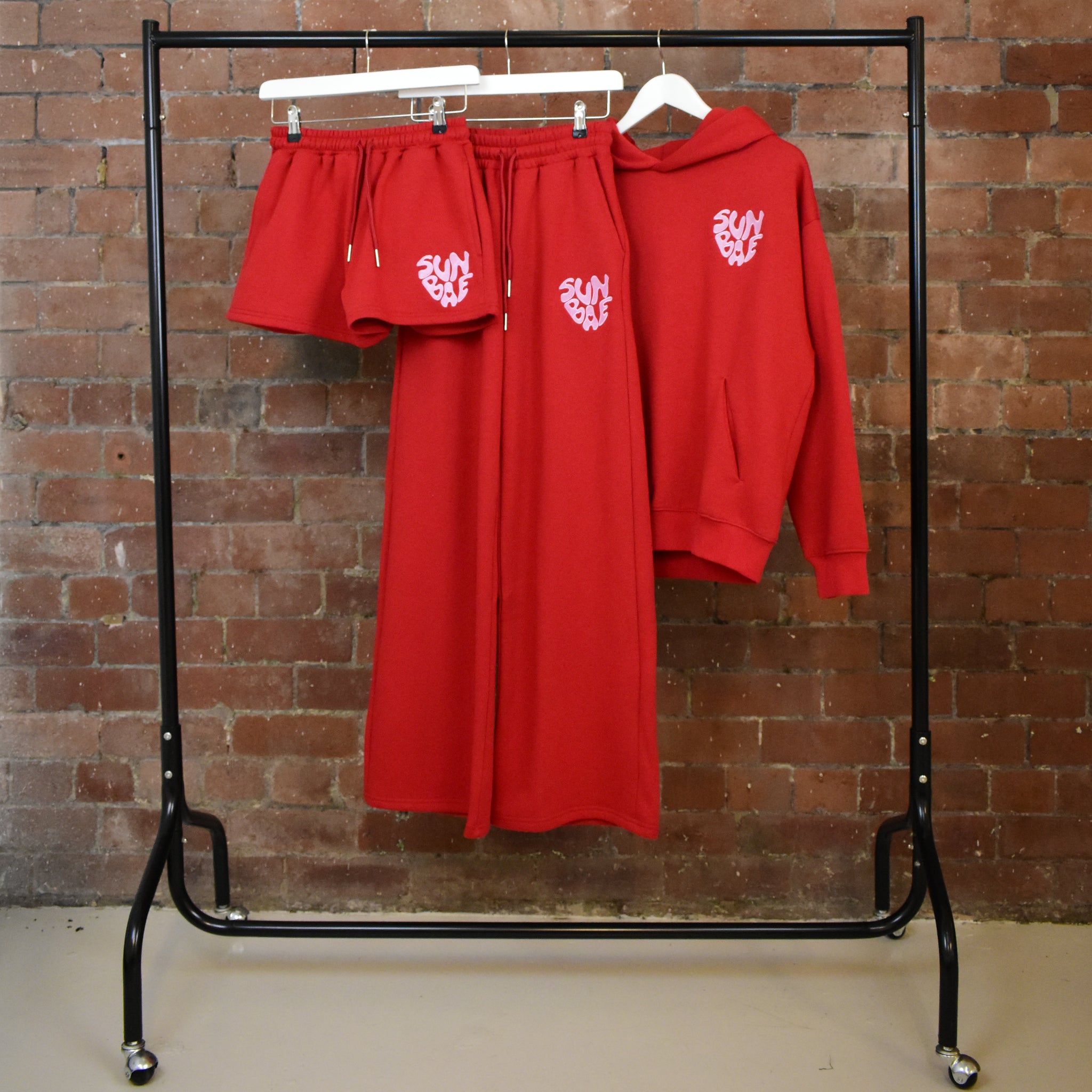 Red SUNBAE Glazed Heart Drop includes hoodie, shorts and sweatpants against a brick wall.