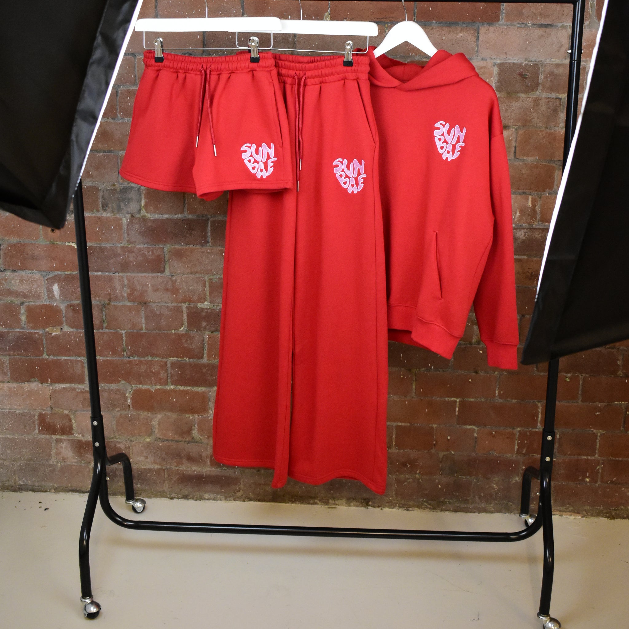 Red SUNBAE Glazed Heart Collection including sweatshirt, shorts, and sweat pants on a garment rack against a brick wall.