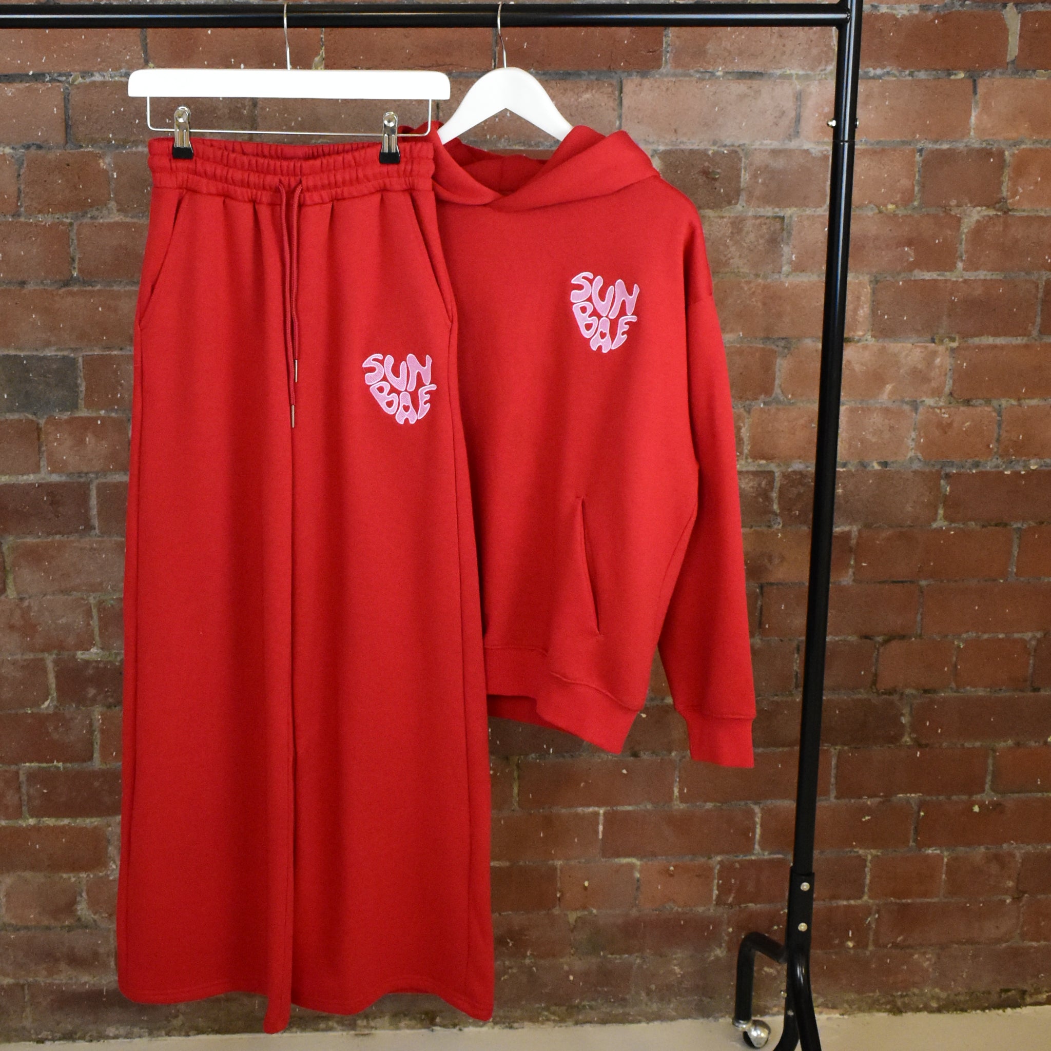 Red SUNBAE hoodie and sweatpants set on a hanger against a brick wall.