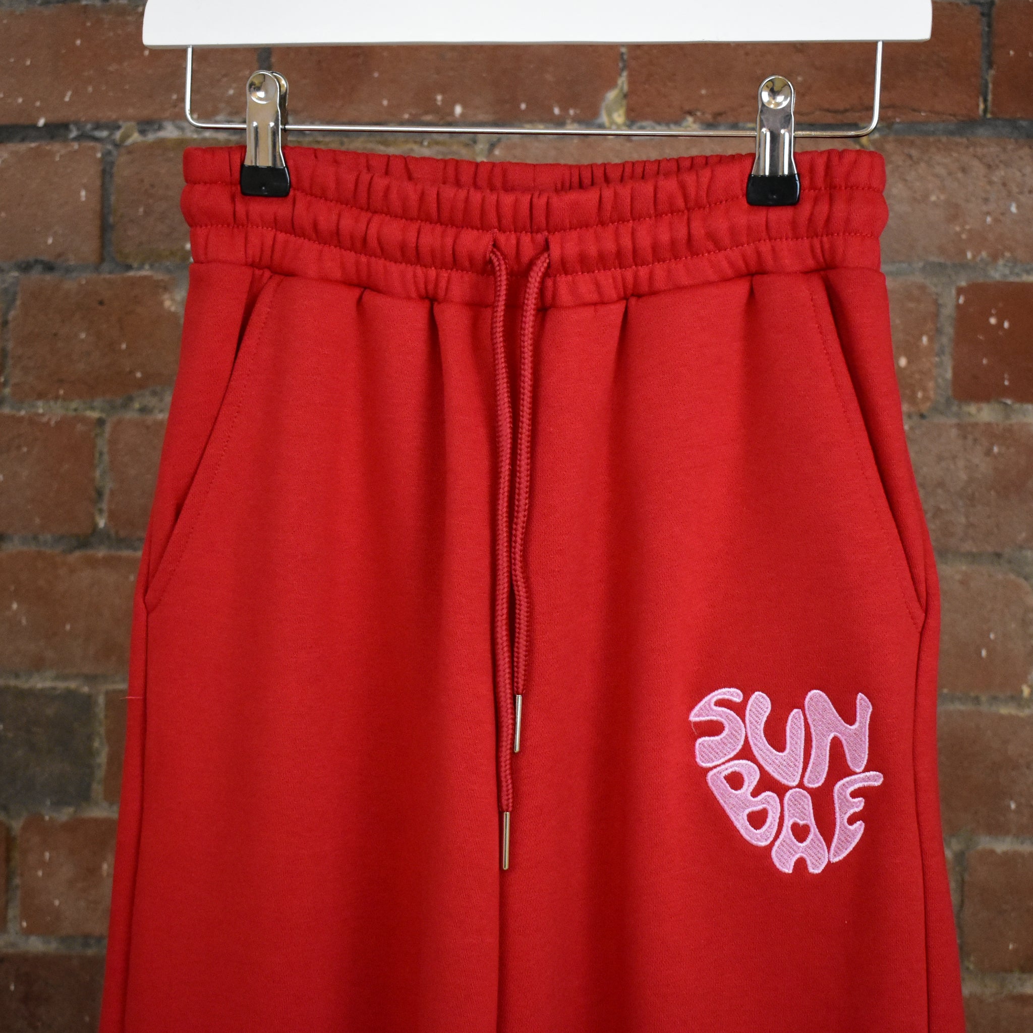 Red SUNBAE sweatpants on a hanger against a brick wall.