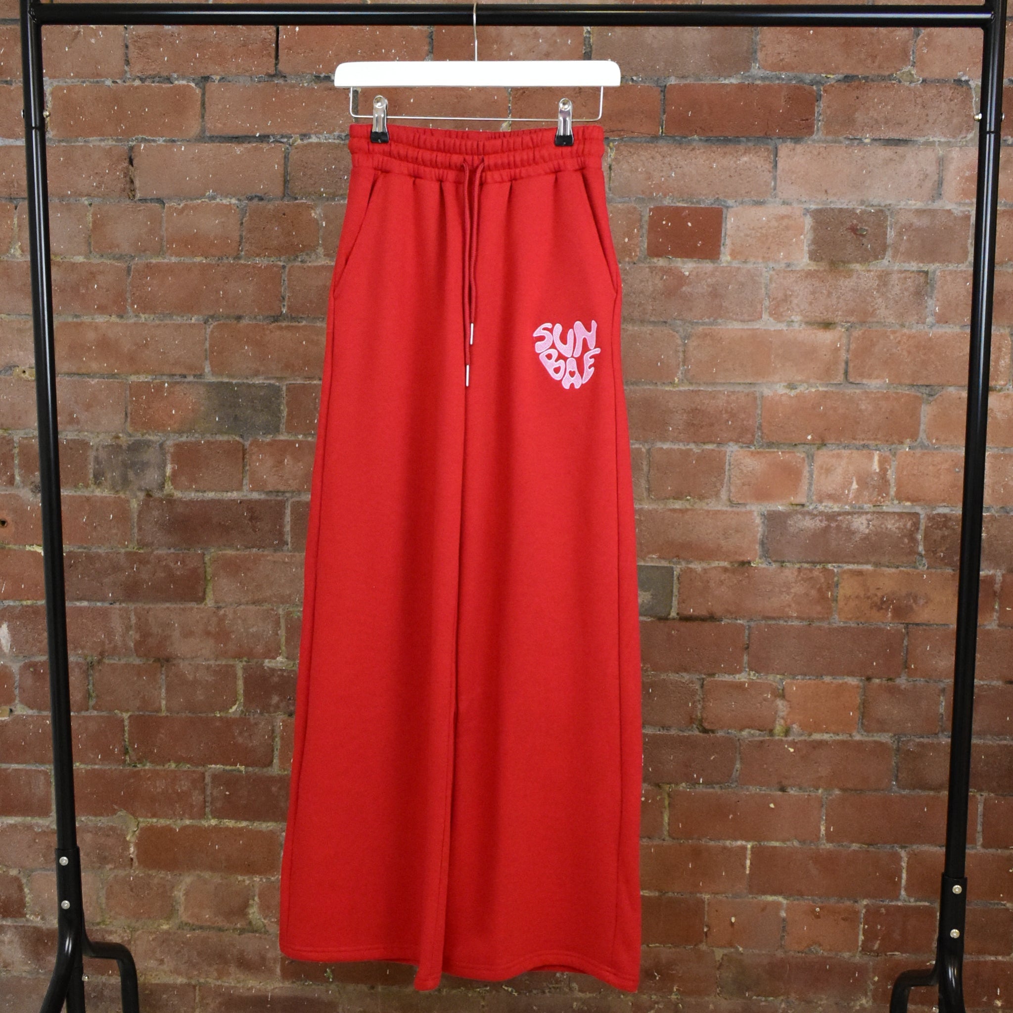 Red SUNBAE sweatpants with a logo on a hanger against a brick wall.