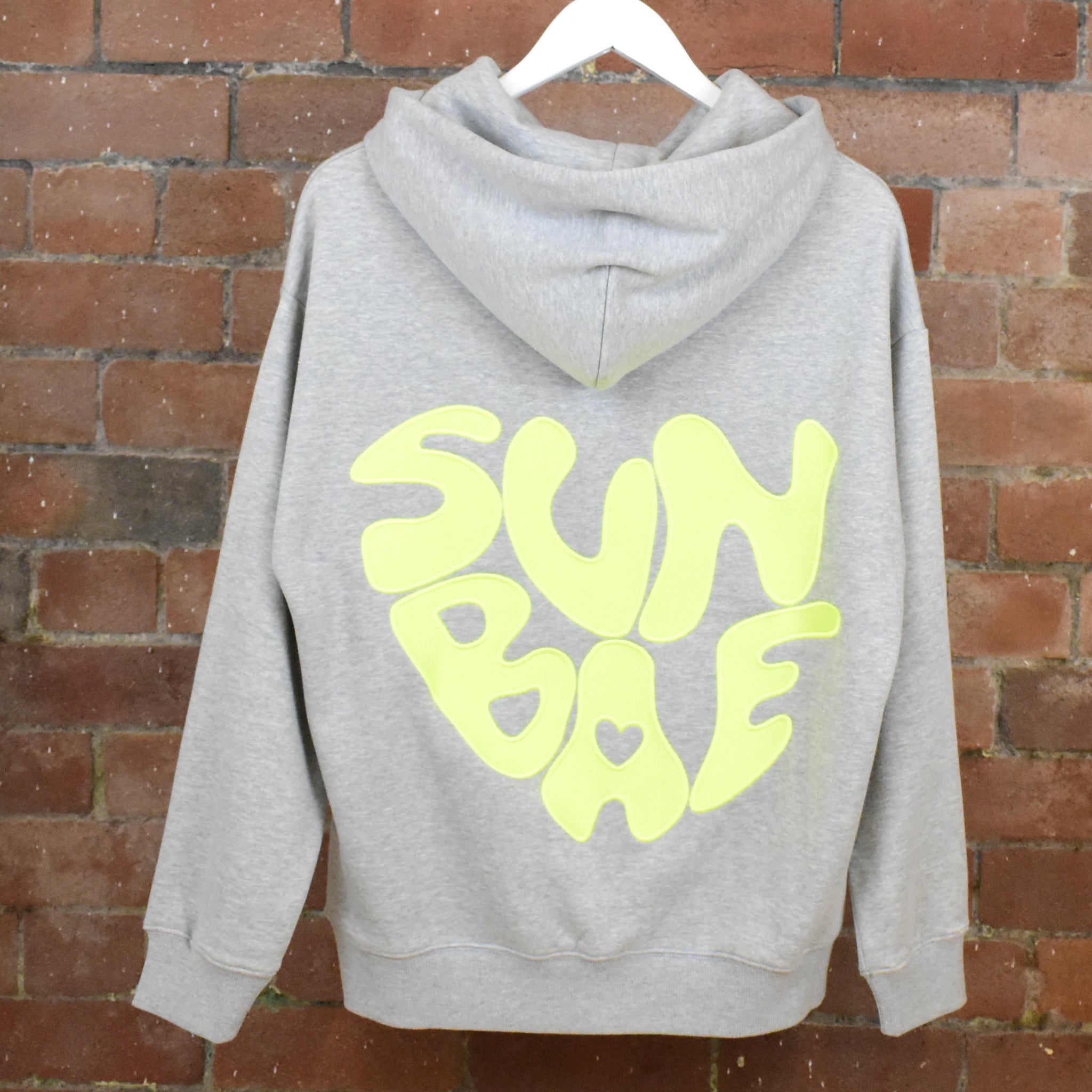 Grey SUNBAE hoodie with logo on the back, hanging against a brick wall.