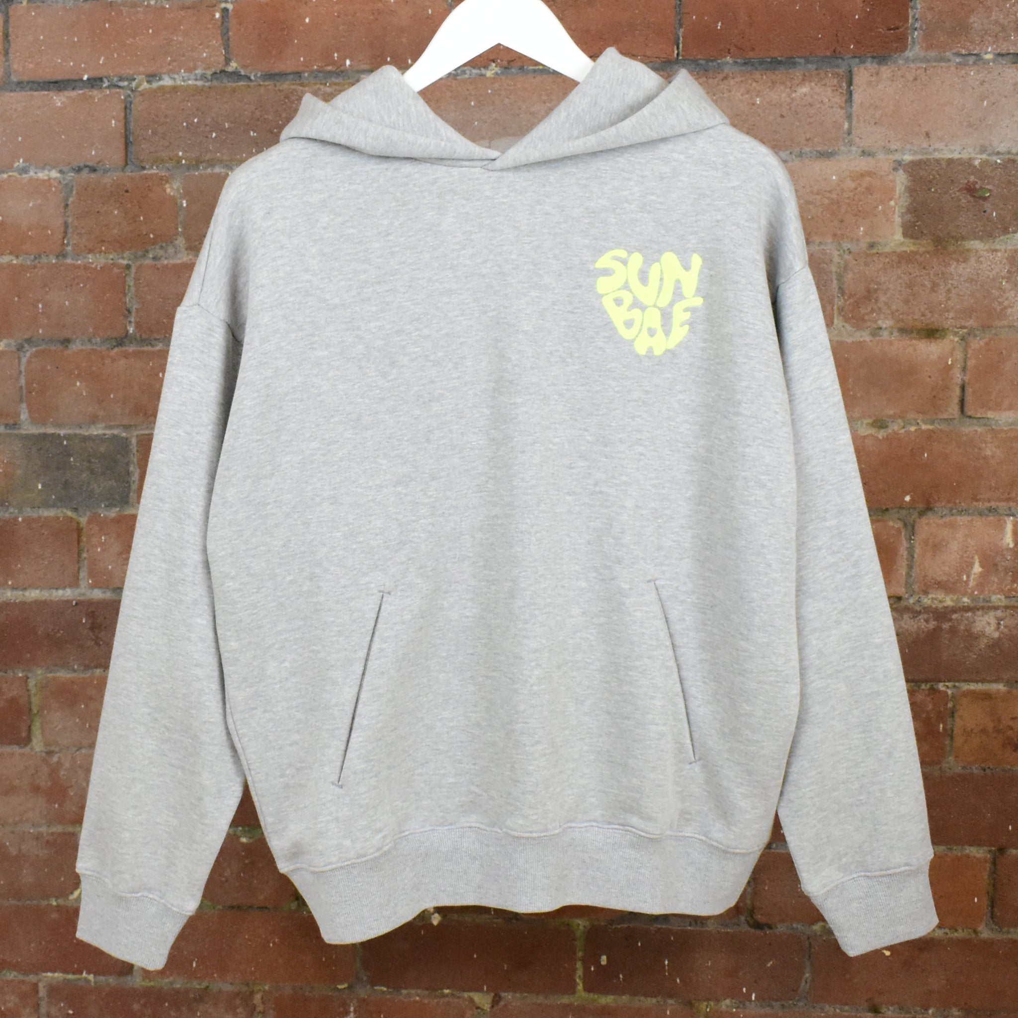 Grey SUNBAE hoodie with a yellow logo on a hanger against a brick wall