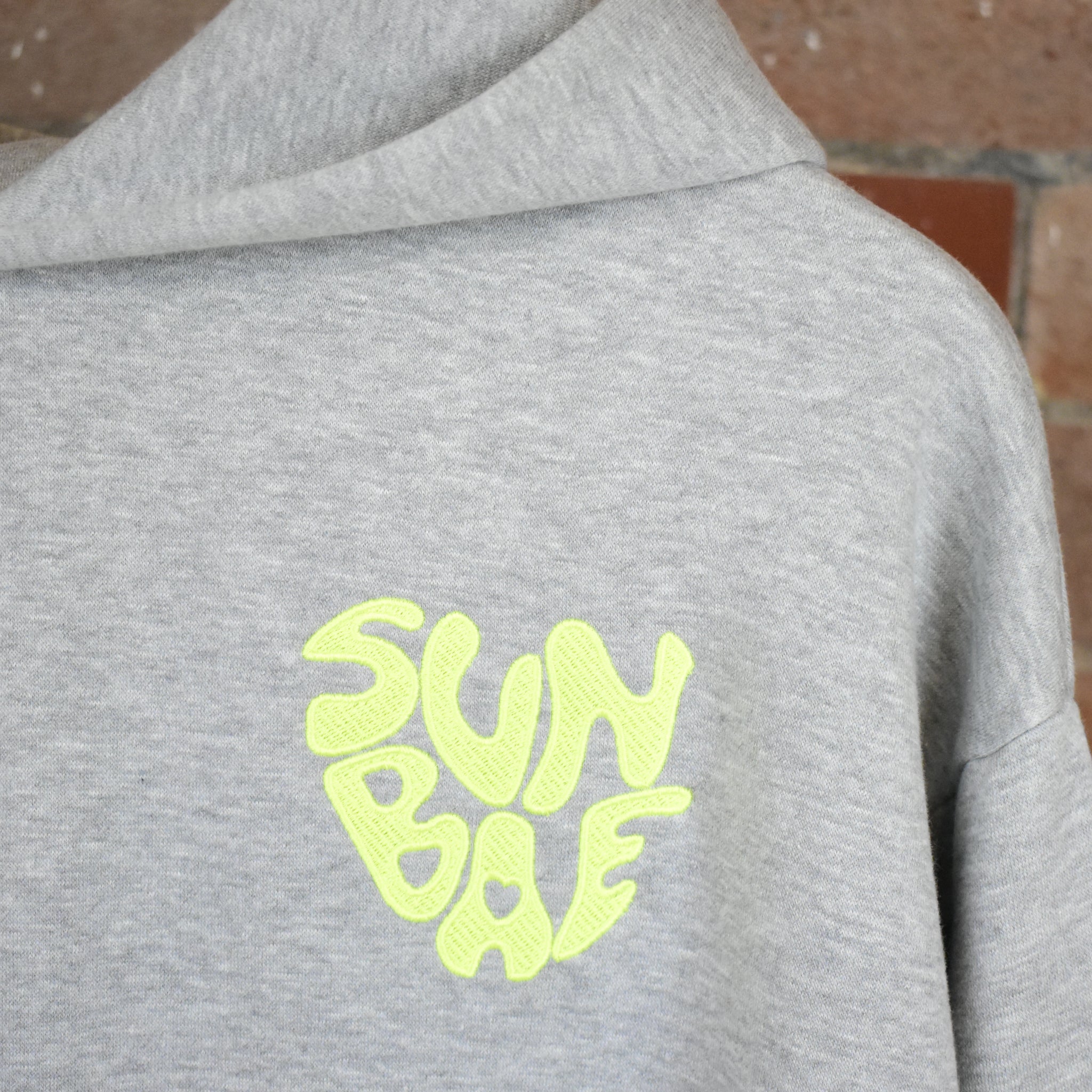 Gray SUNBAE hoodie with logo in neon yellow on a brick wall background