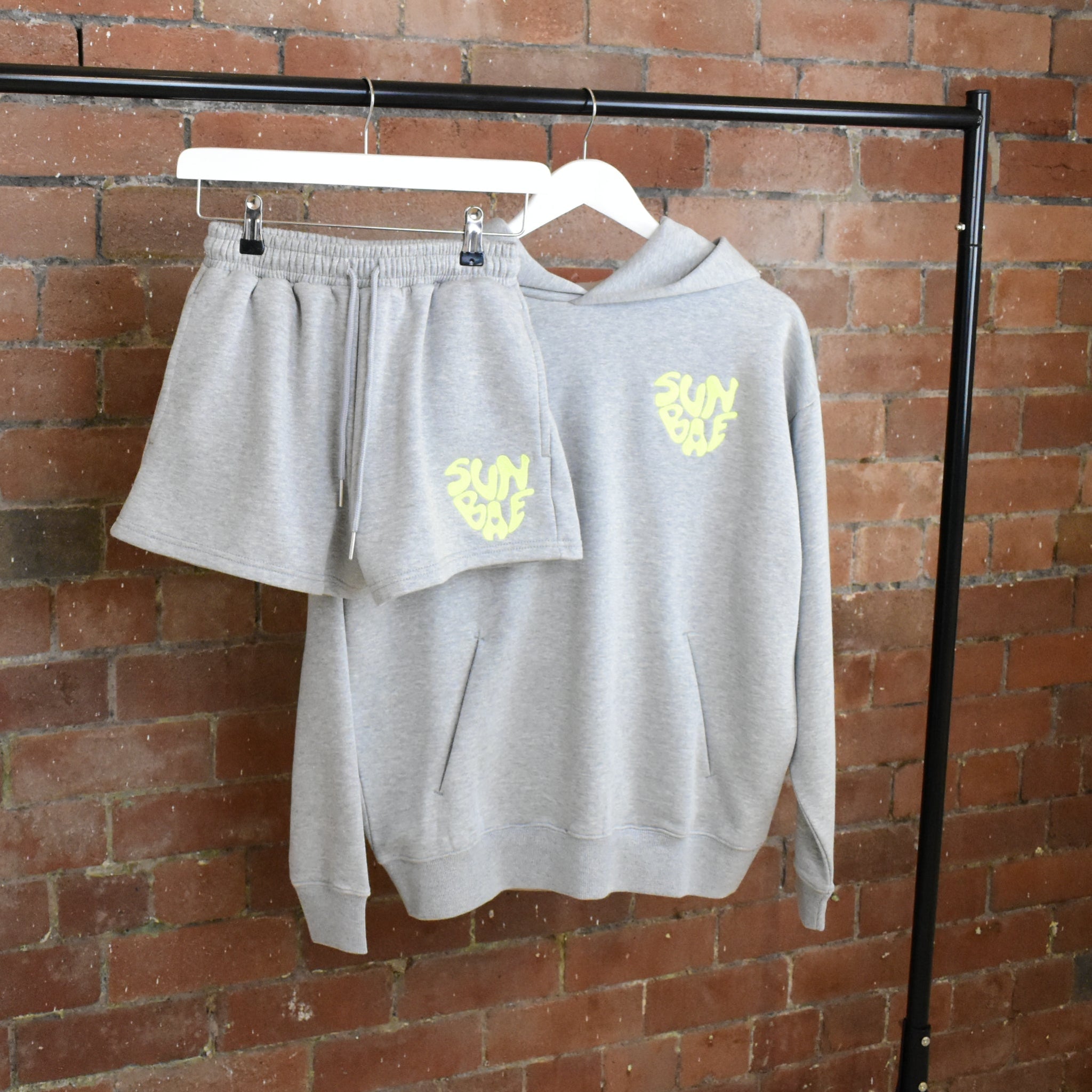 Gray SUNBAE sweatshirt and shorts set with yellow logo on hangers against a brick wall.