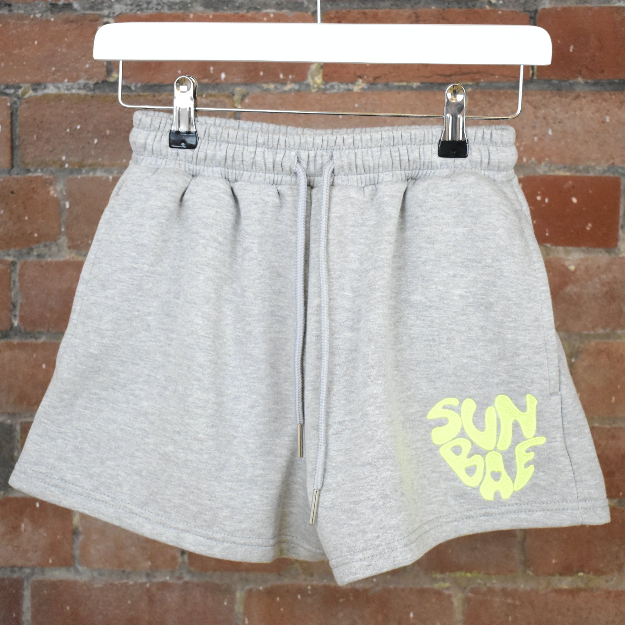 Grey SUNBAE shorts on a hanger against a brick wall.