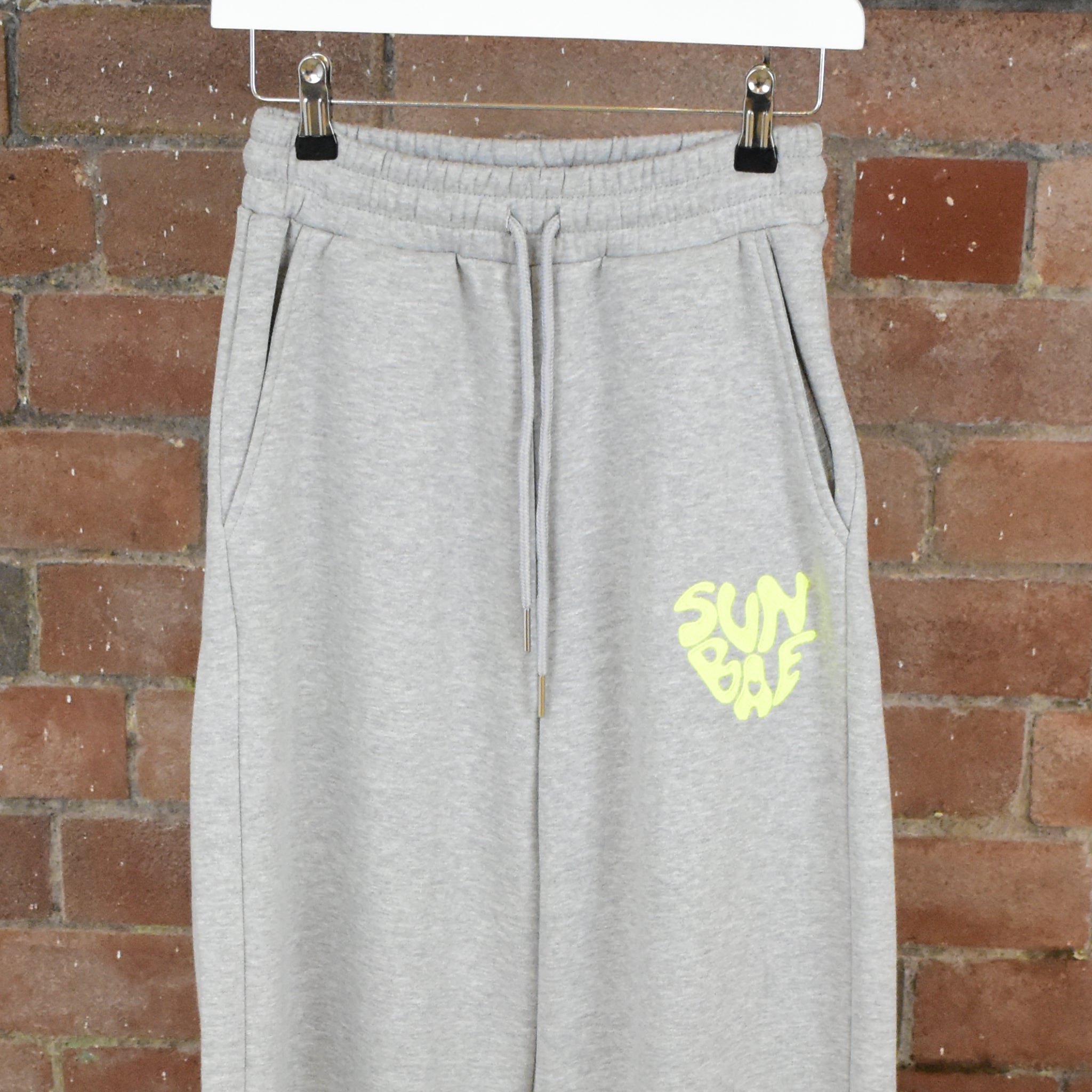 Gray SUNBAE Sweat Pants on a hanger against a brick wall.