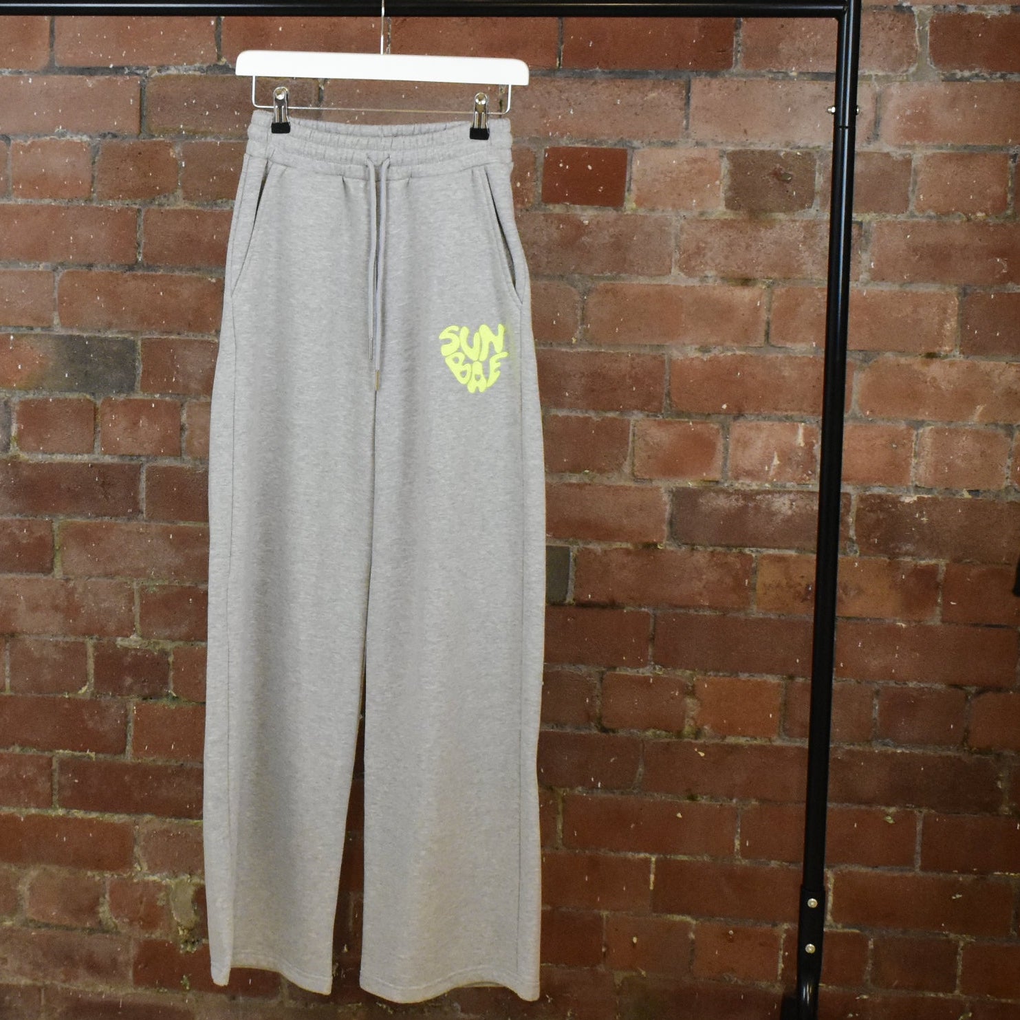 Grey SUNBAE sweatpants with a yellow logo on a hanger against a brick wall.