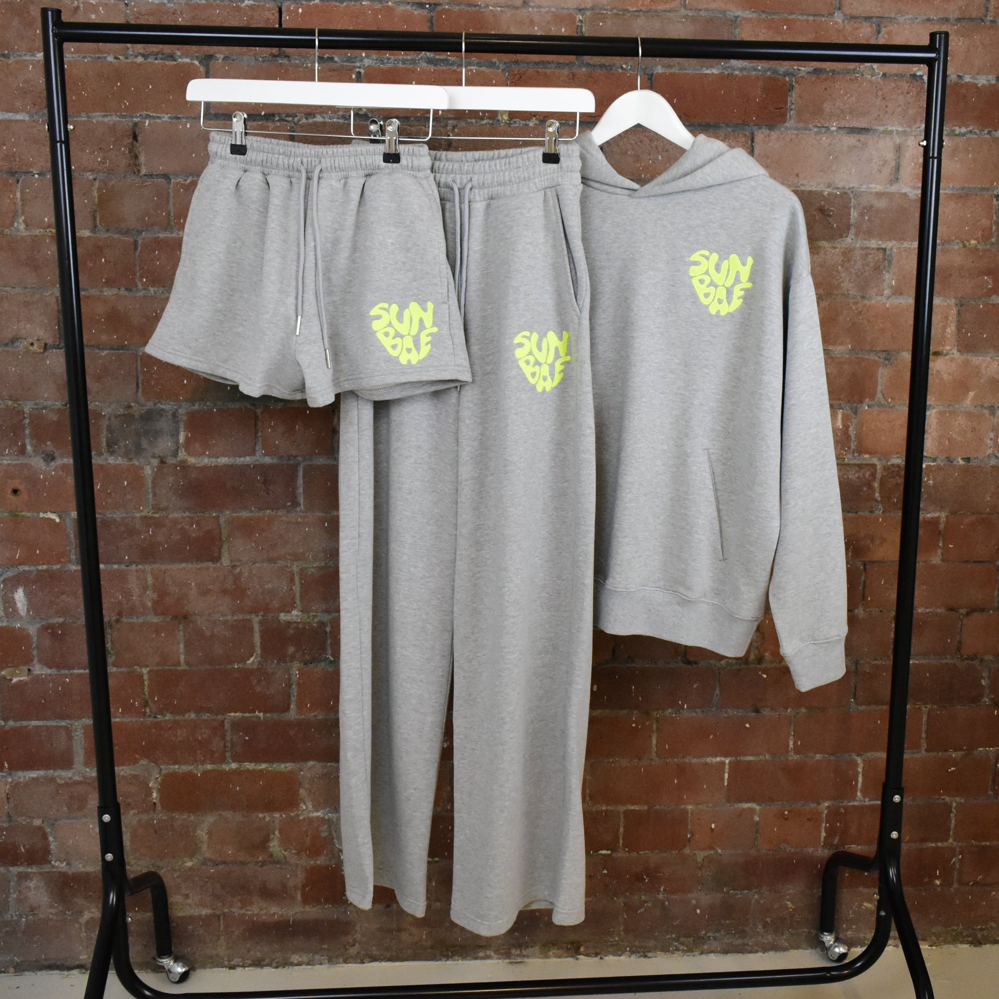 Grey SUNBAE Hoodie, Shorts, and Sweat Pants on hangers against a brick wall.