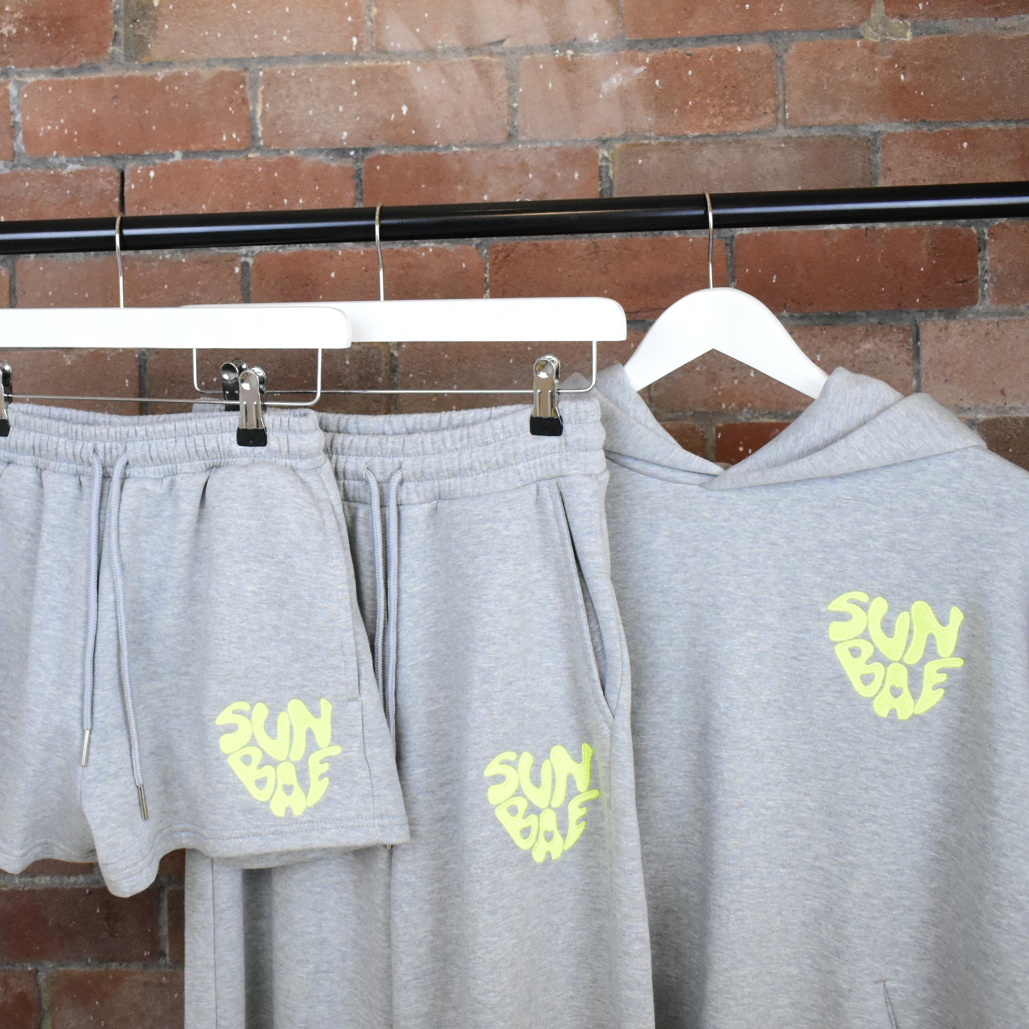 Grey SUNBAE Sweat Pants and Hoodie on hangers against a brick wall.