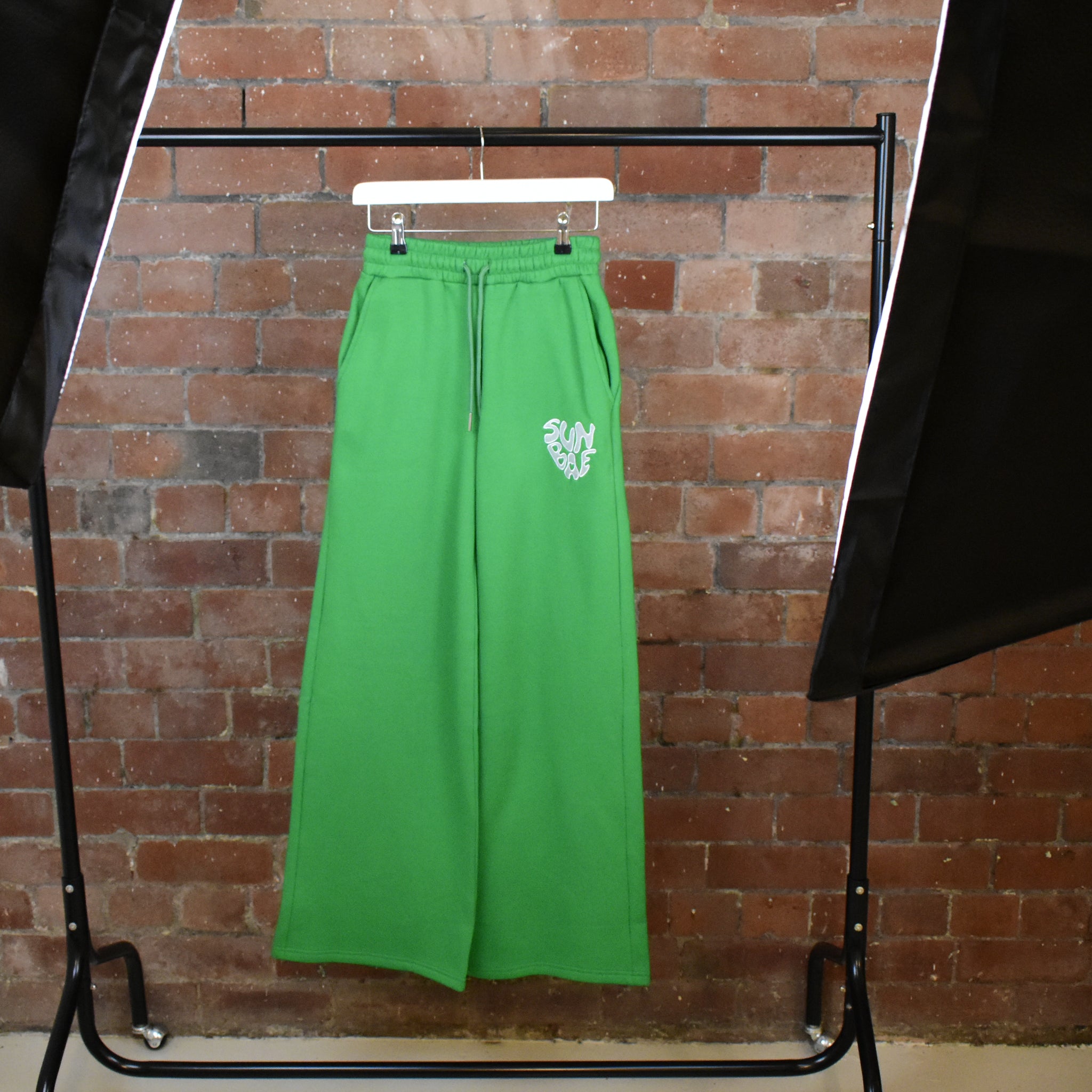 Green SUNBAE Sweat Pants on a hanger against a brick wall