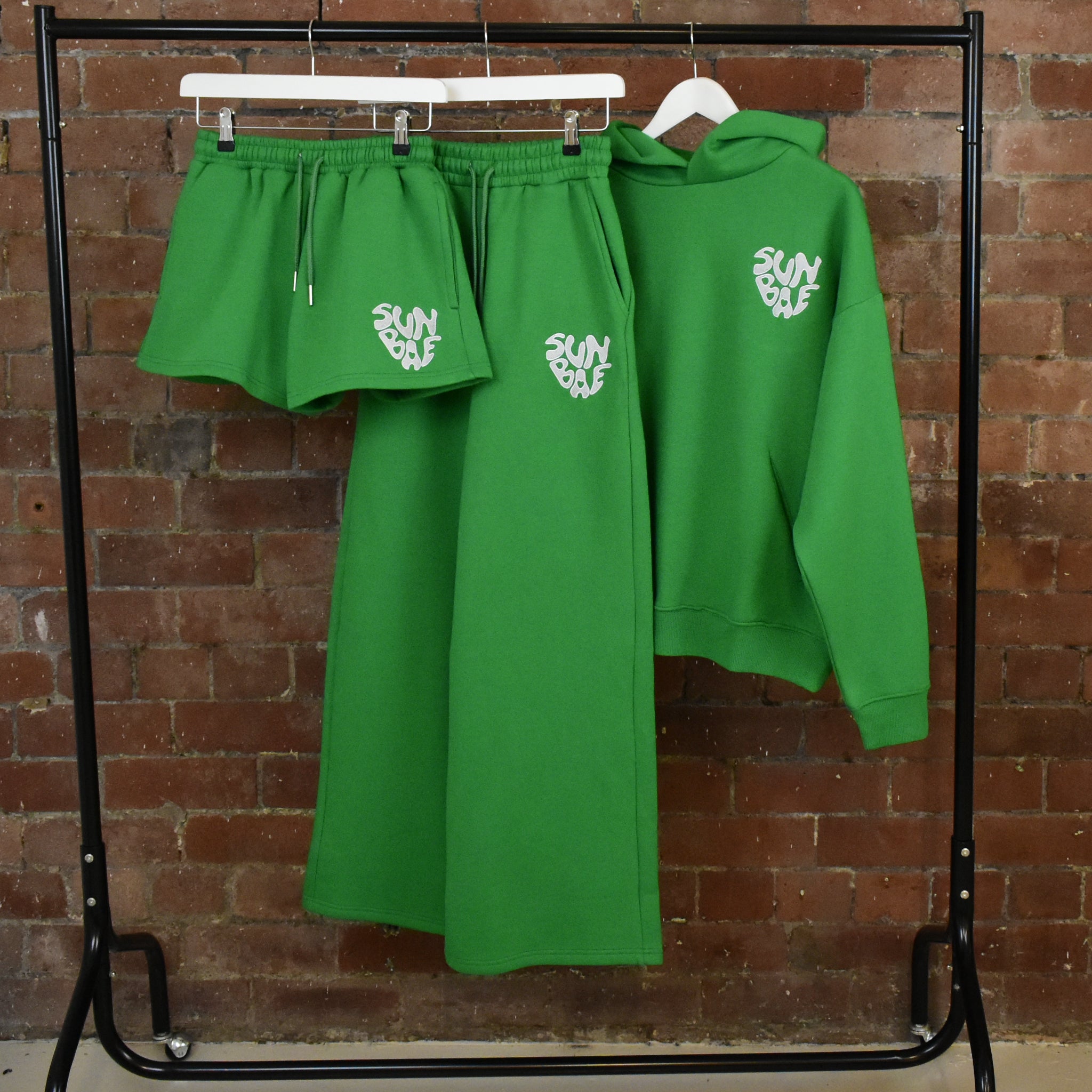 Green SUNBAE hoodie, sweatpants, and shorts set with logo on a rack against a brick wall