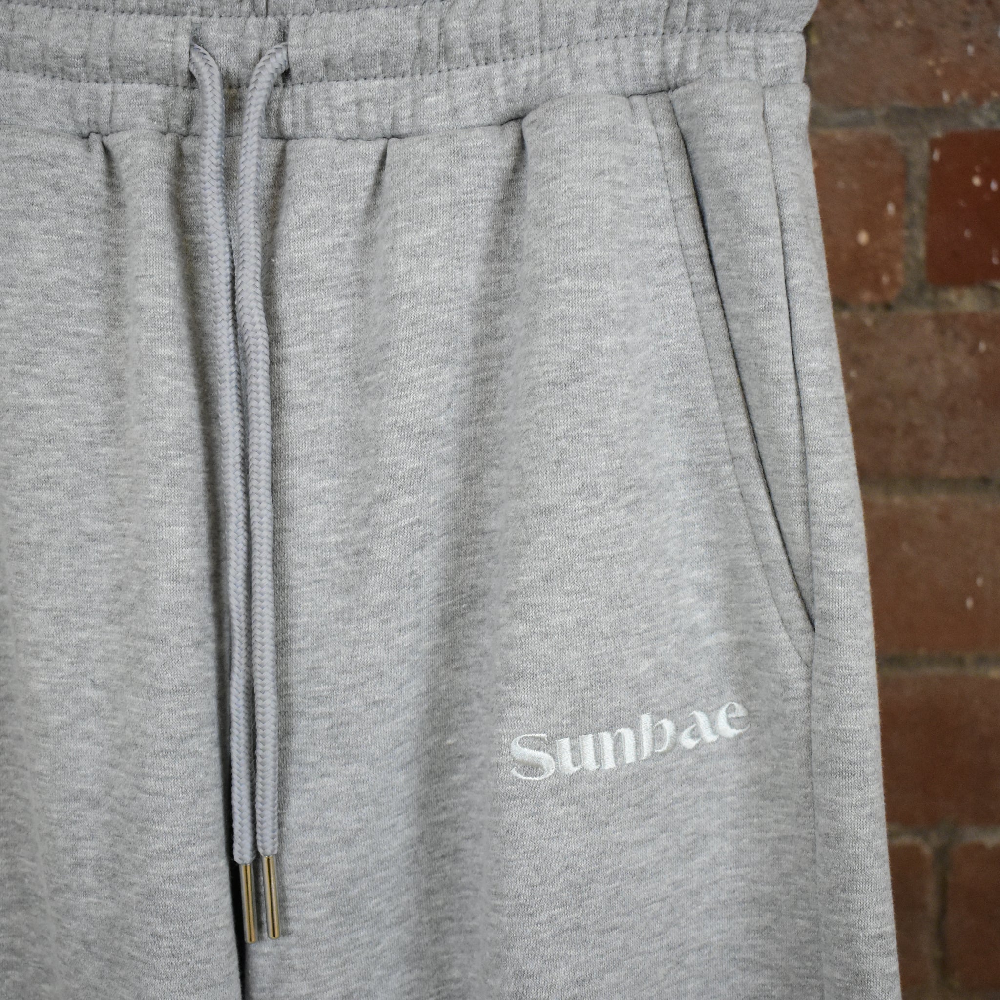 Grey SUNBAE sweatpants with logo against a brick wall