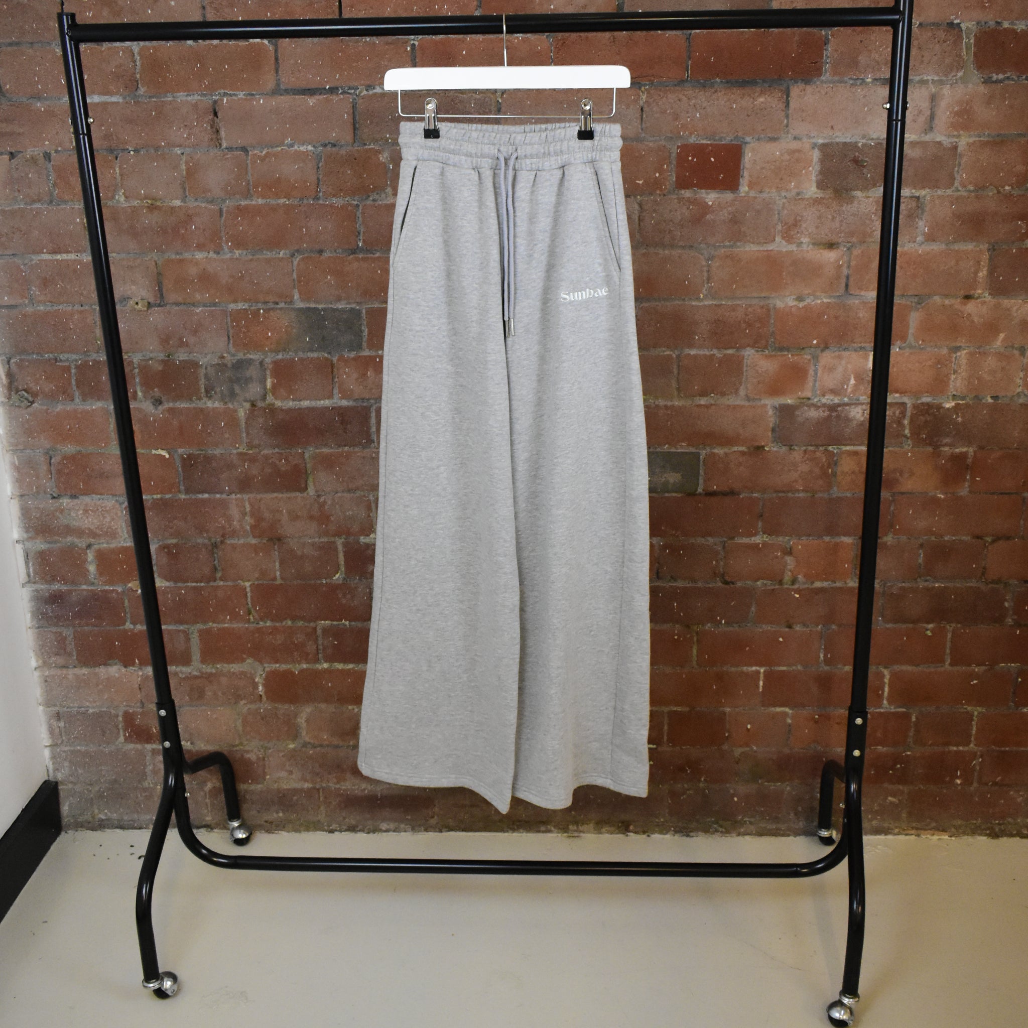 Grey SUNBAE sweatpants hanging on a black garment rack against a brick wall.