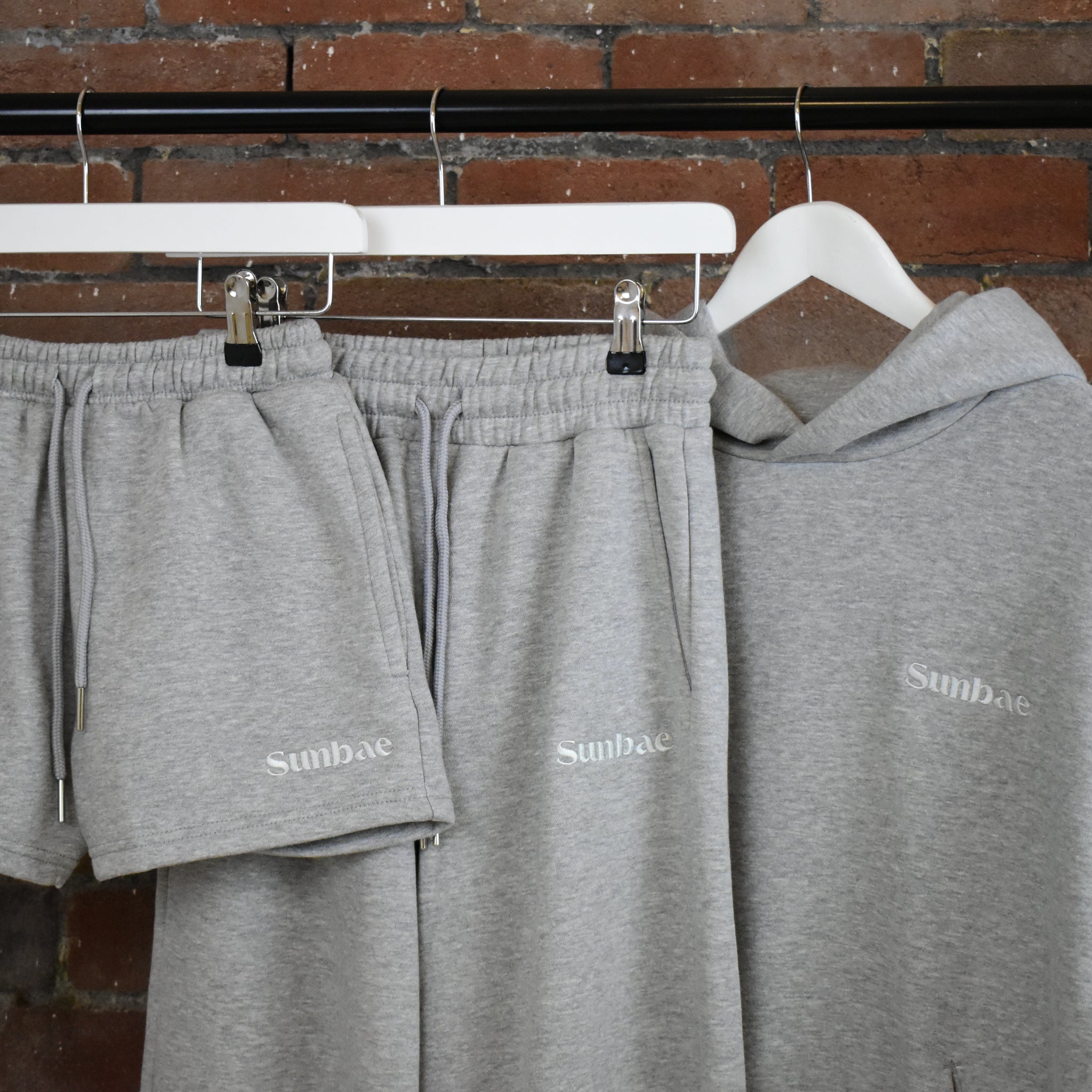 Grey SUNBAE sweatpants and hoodie set on hangers against a brick wall.