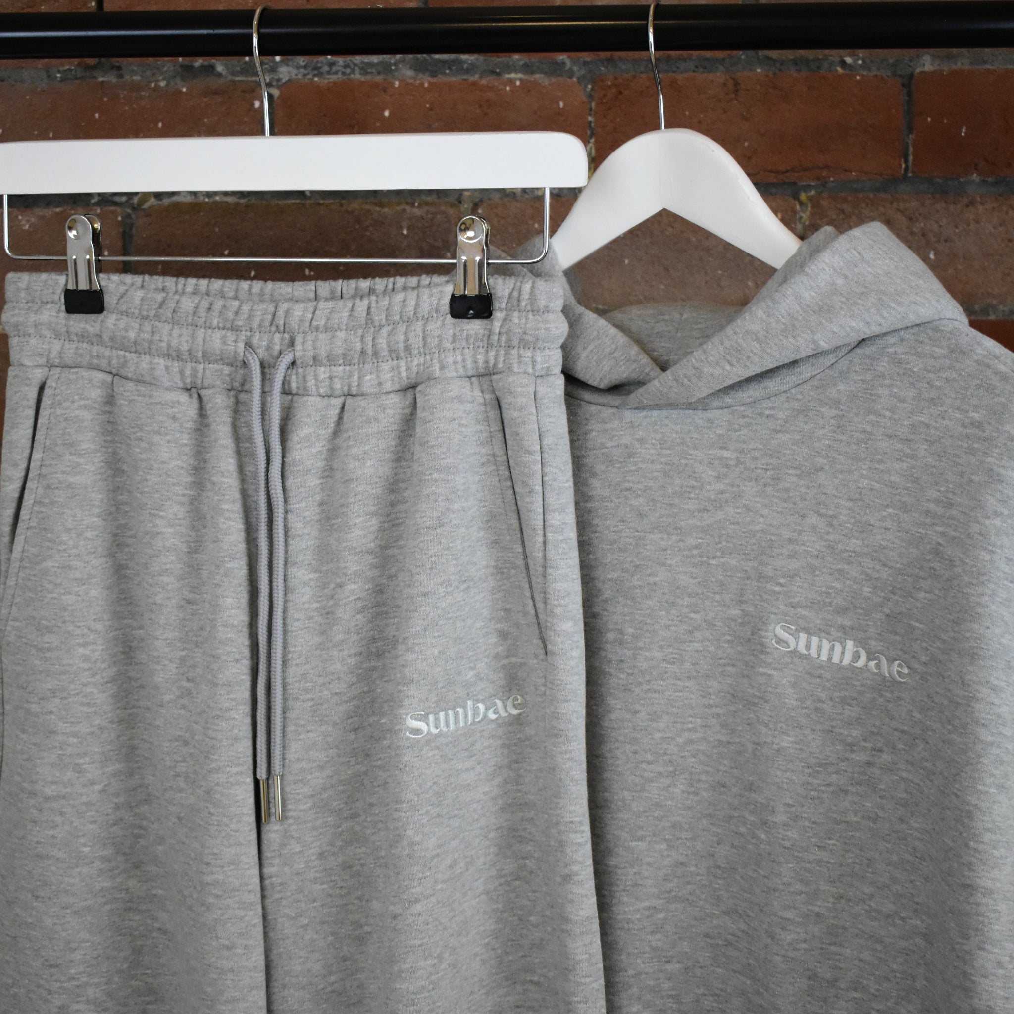Grey SUNBAE hoodie and sweatpants on hangers against a brick wall.