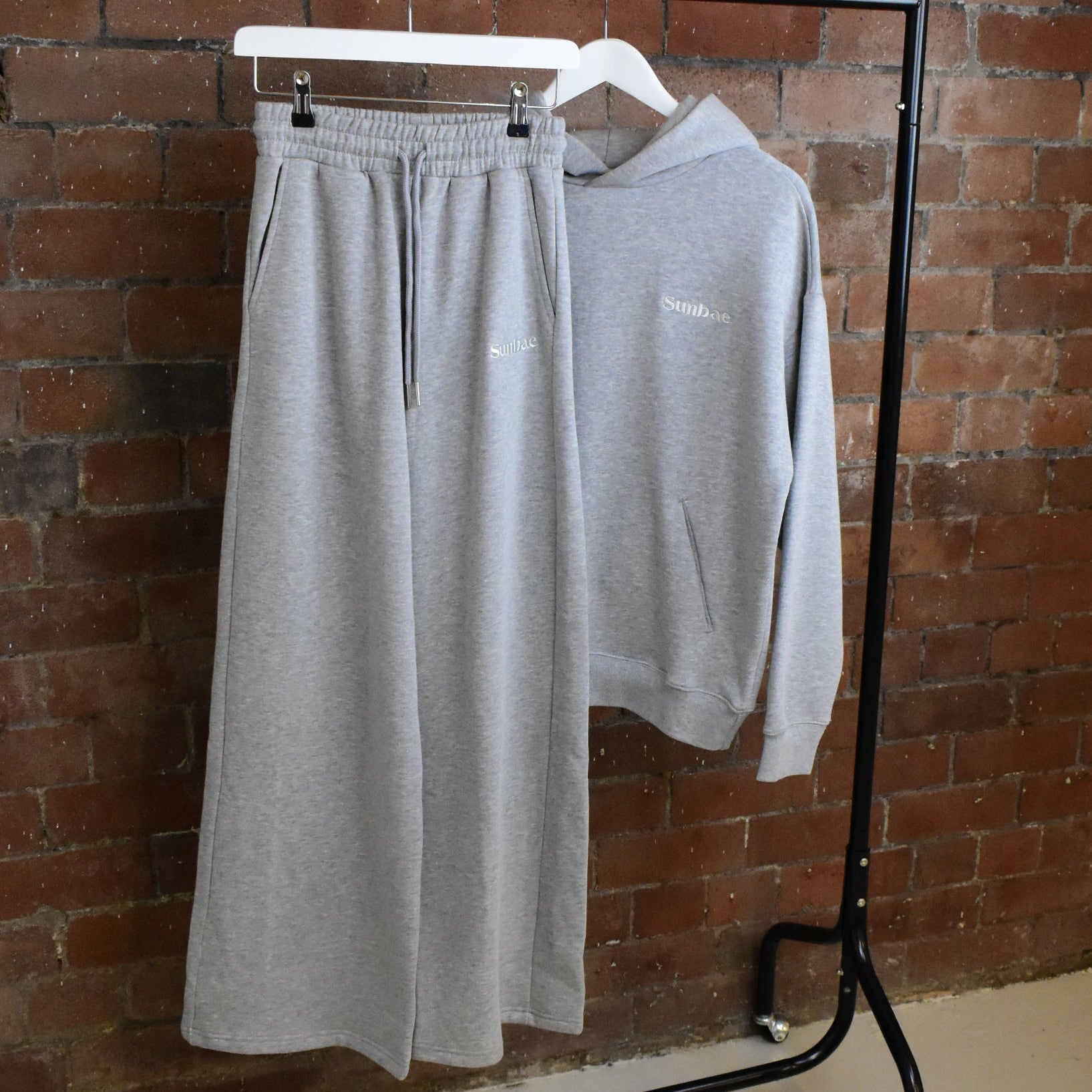 Grey SUNBAE sweatpants and hoodie on a black coat rack against a brick wall.