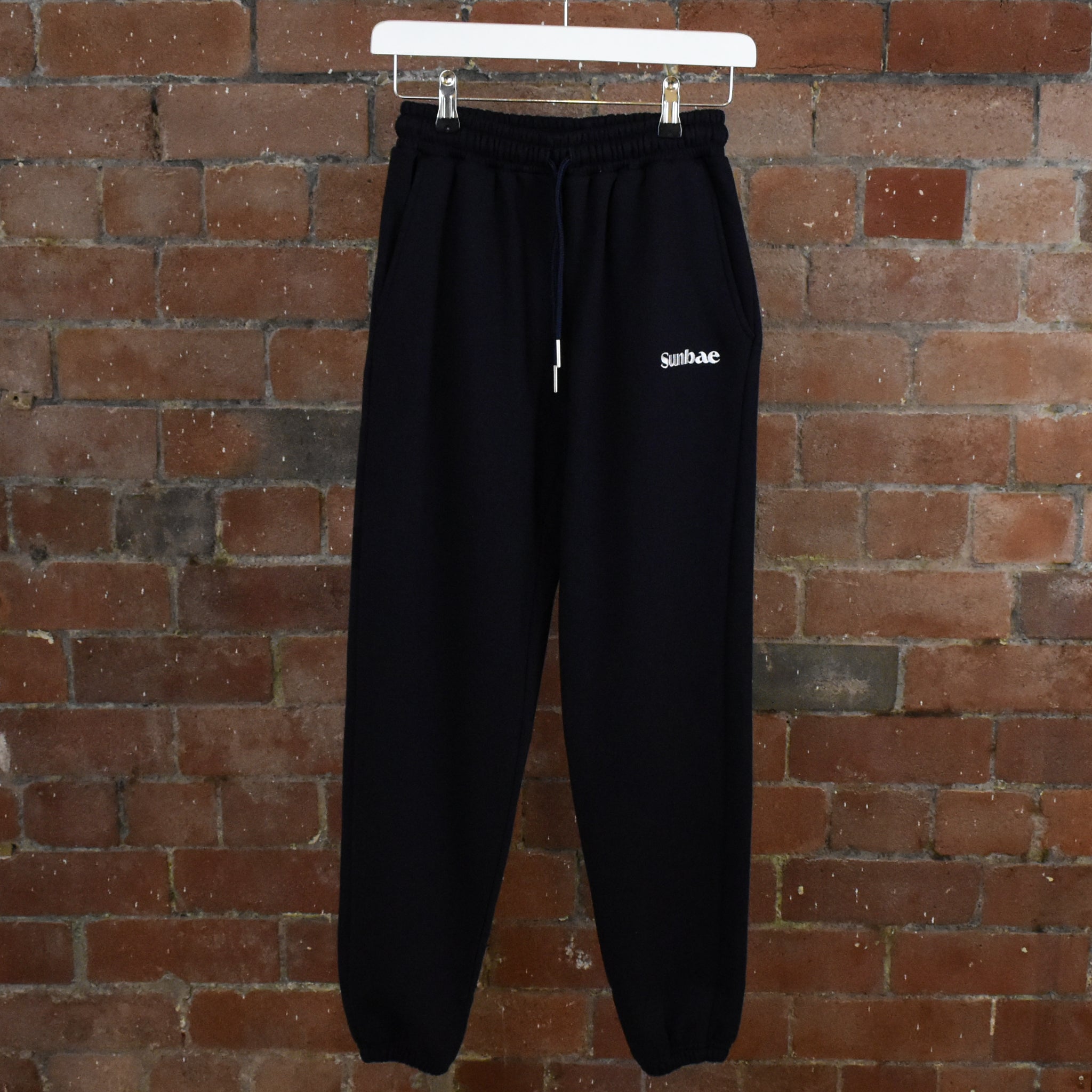 Navy SUNBAE sweatpants with a logo on a hanger against a brick wall