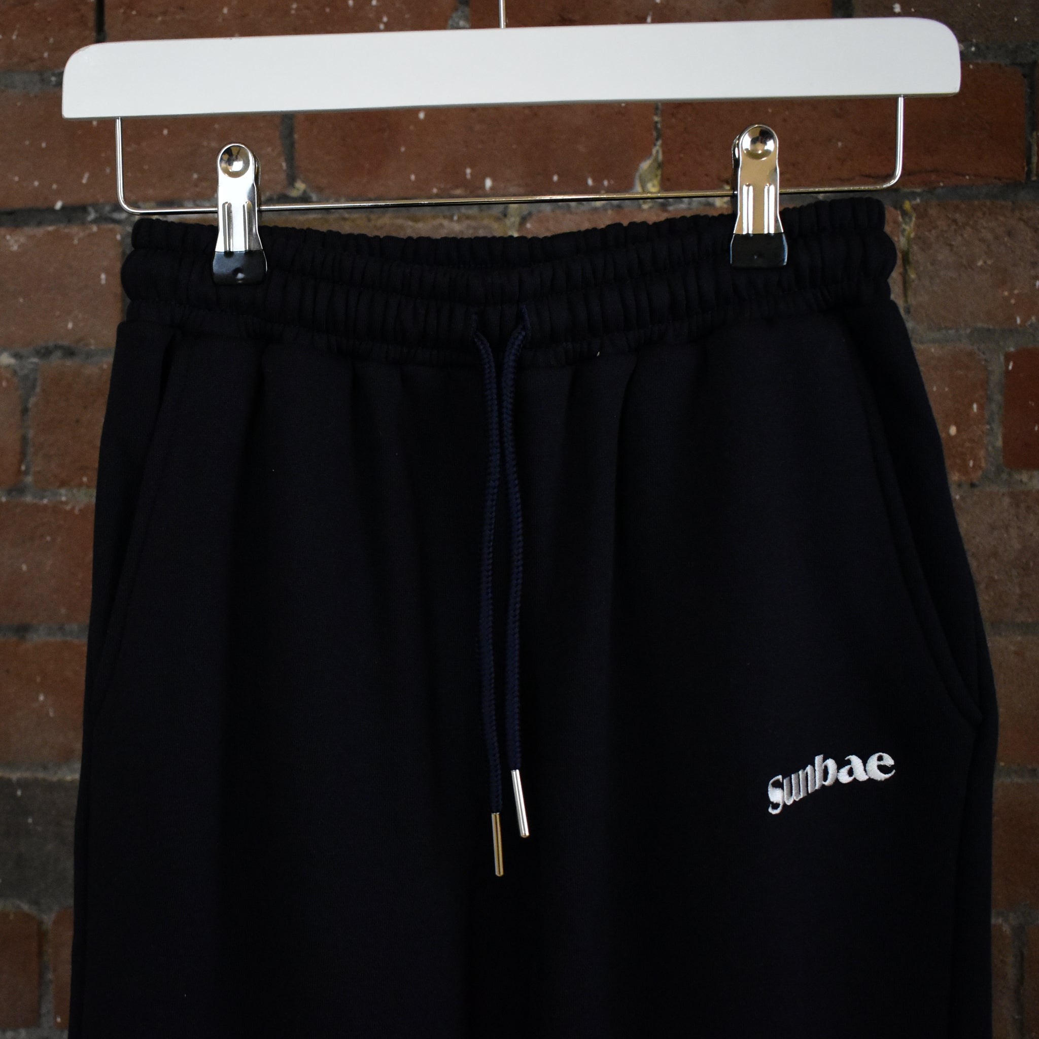Navy SUNBAE Sweat Pants with branding on a hanger against a brick wall.