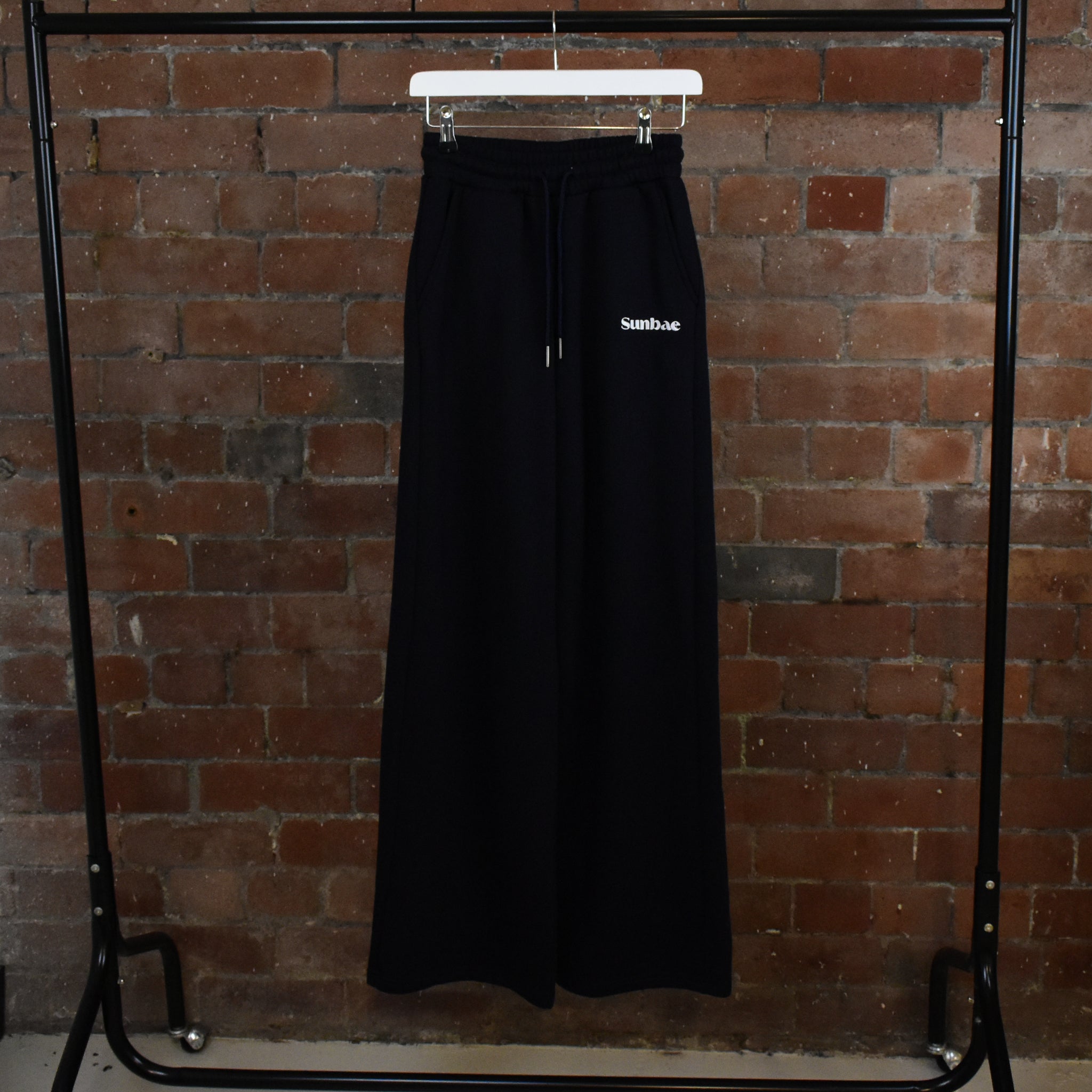 Navy Sweayt Pant with 'SUNBAE' branding on a hanger against a brick wall.