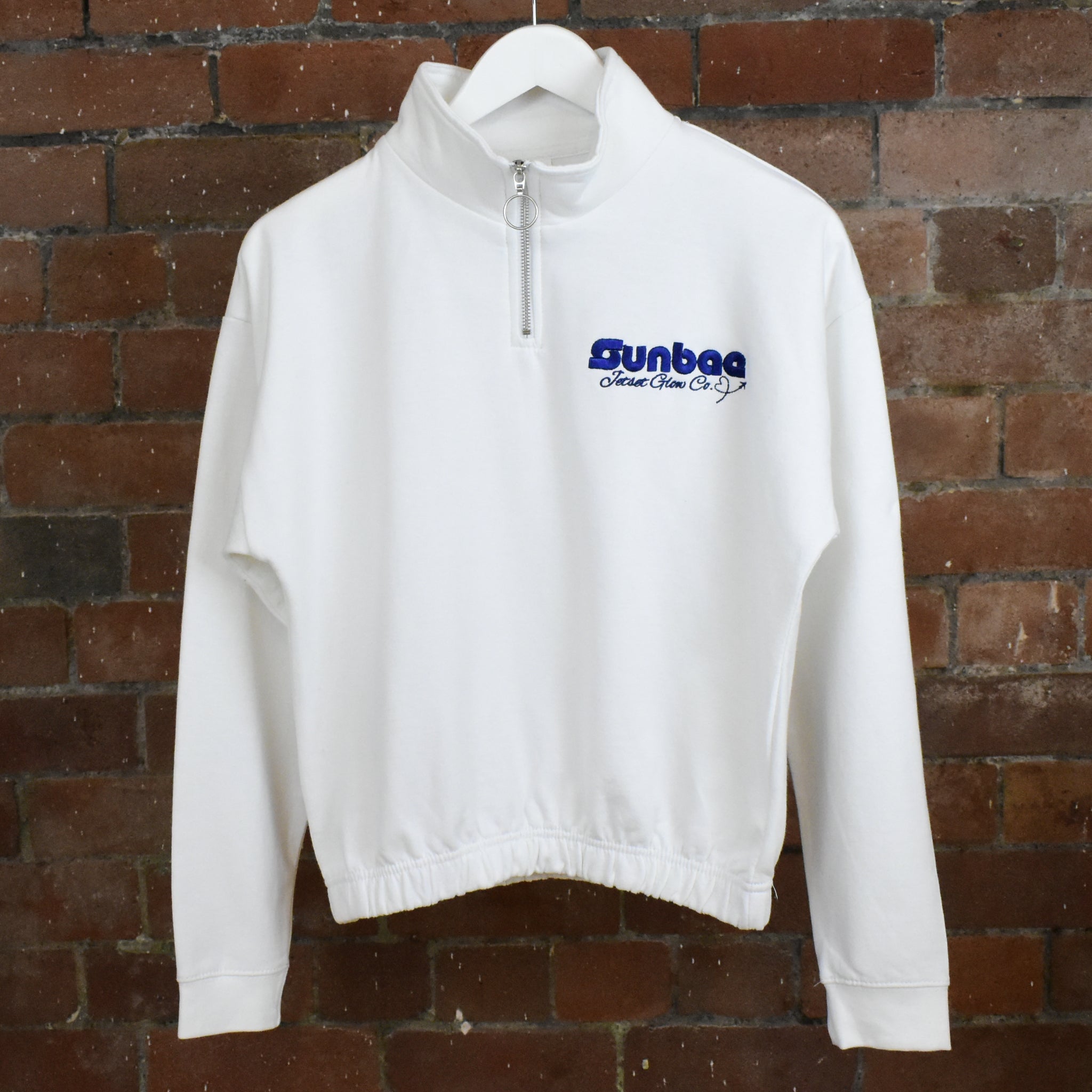 White SUNBAE half-zip sweatshirt with blue logo on a hanger against a brick wall.