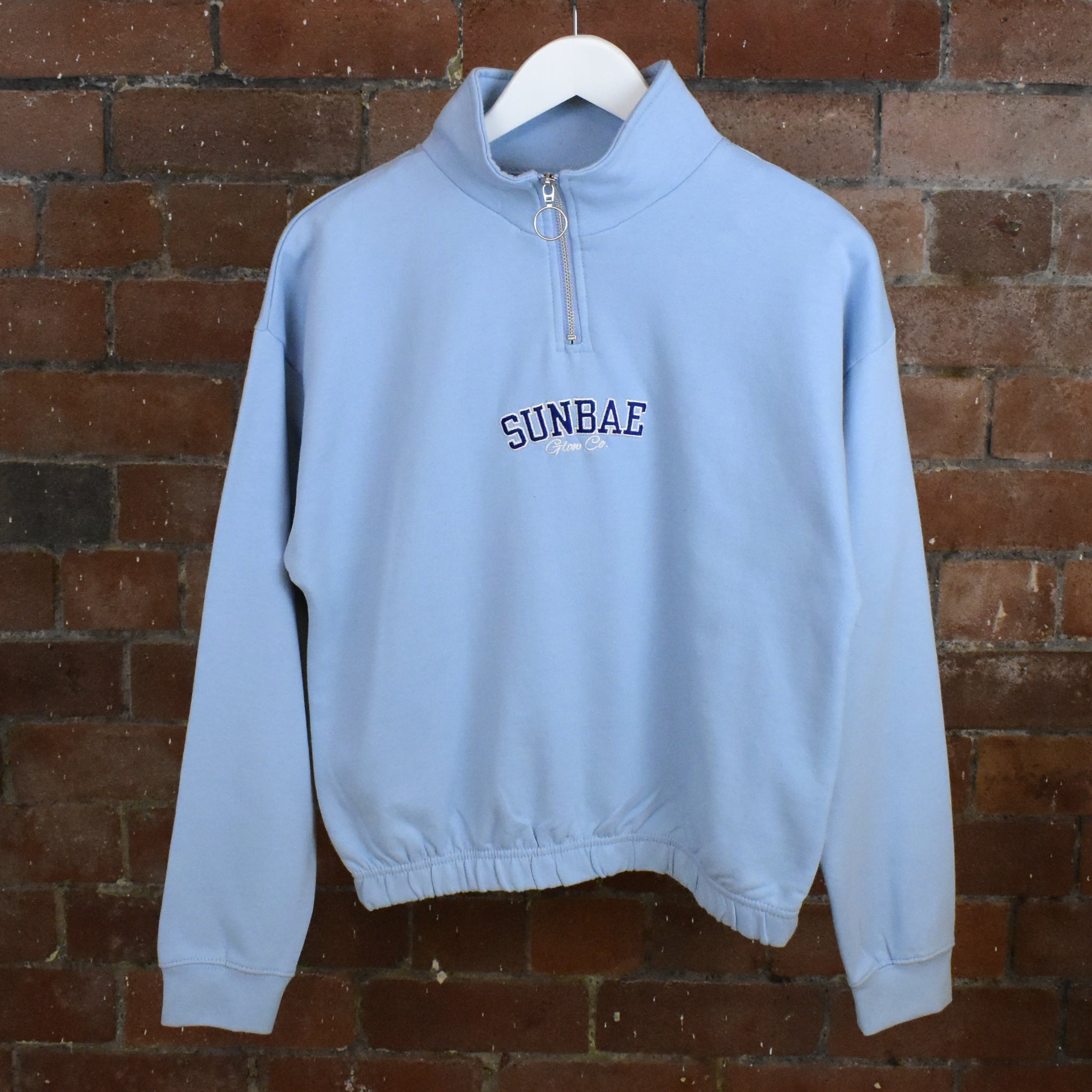 Light blue sweatshirt with 'SUNBAE' text on a hanger against a brick wall.
