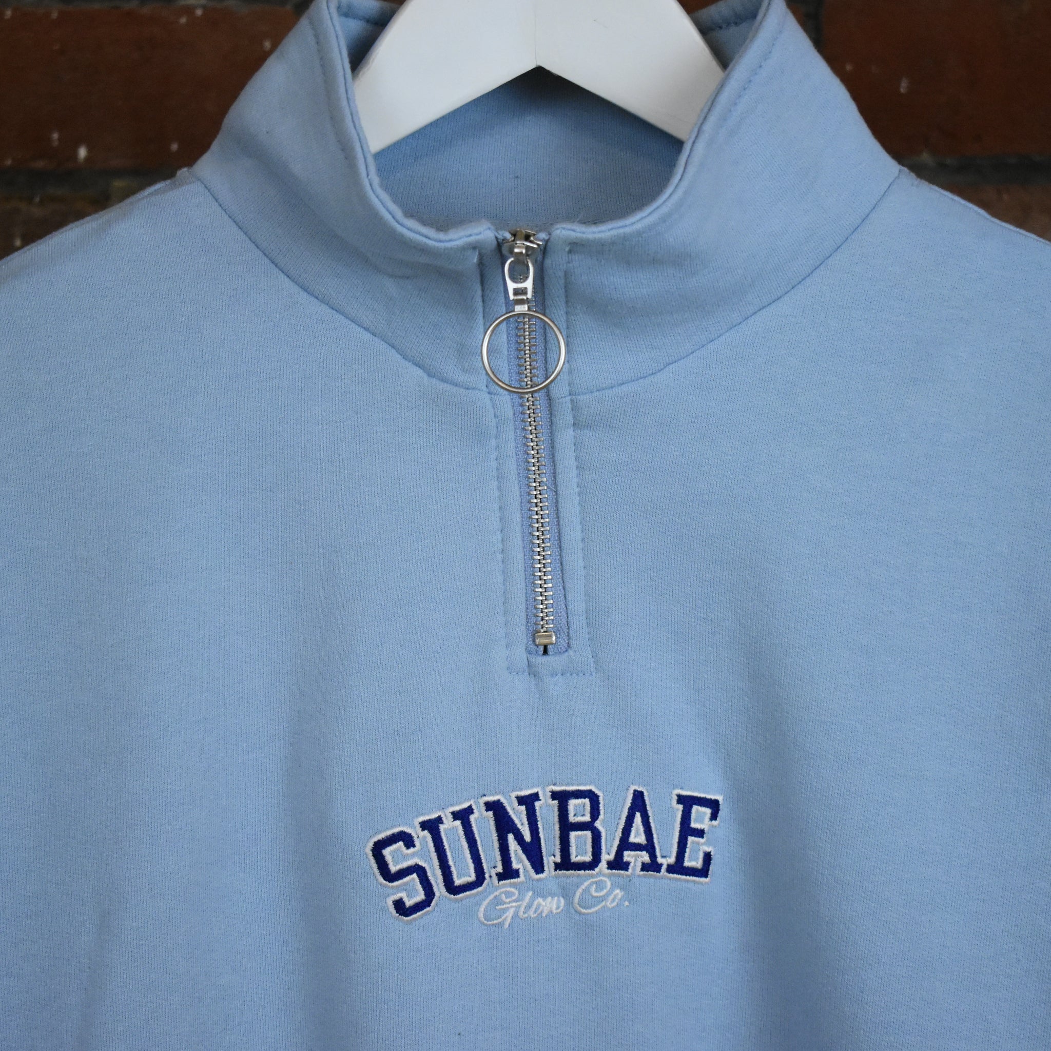 Blue pullover with a zipper and 'SUNBAE' branding on a hanger.