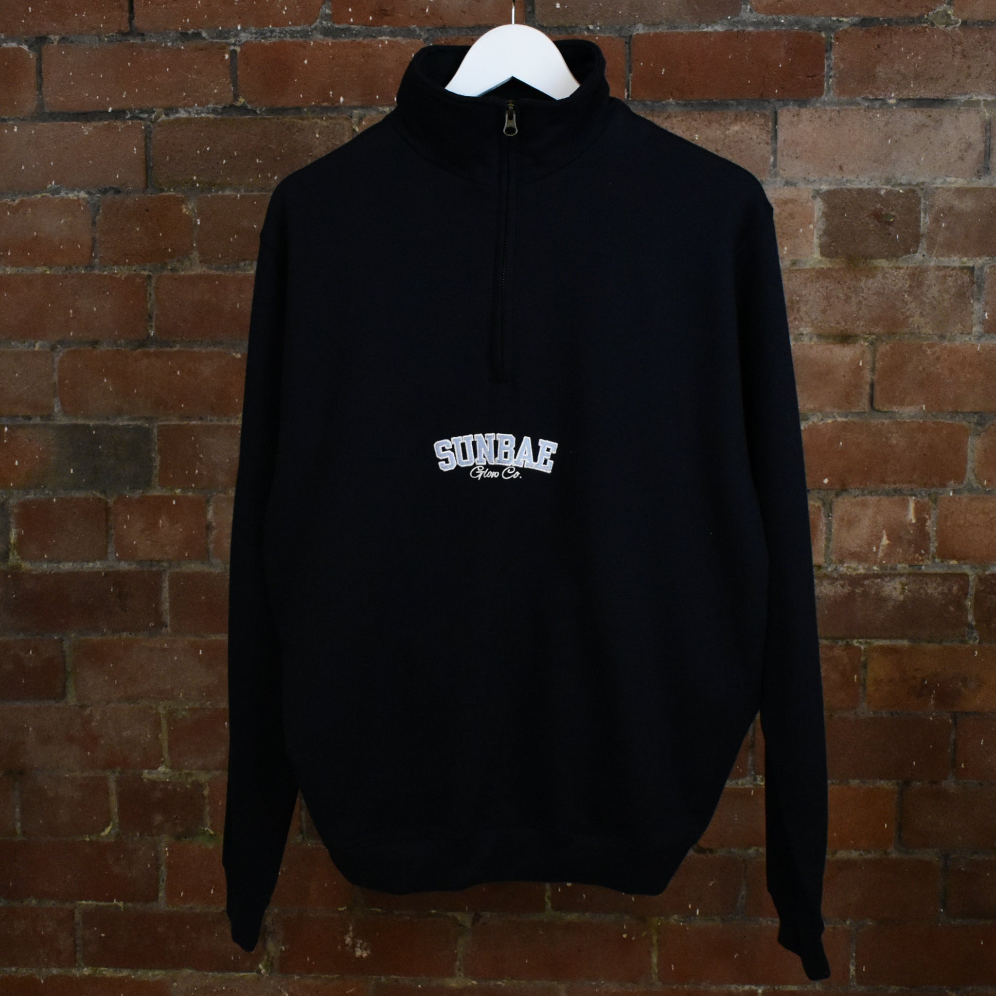 Navy SUNBAE hoodie with logo on a hanger against a brick wall