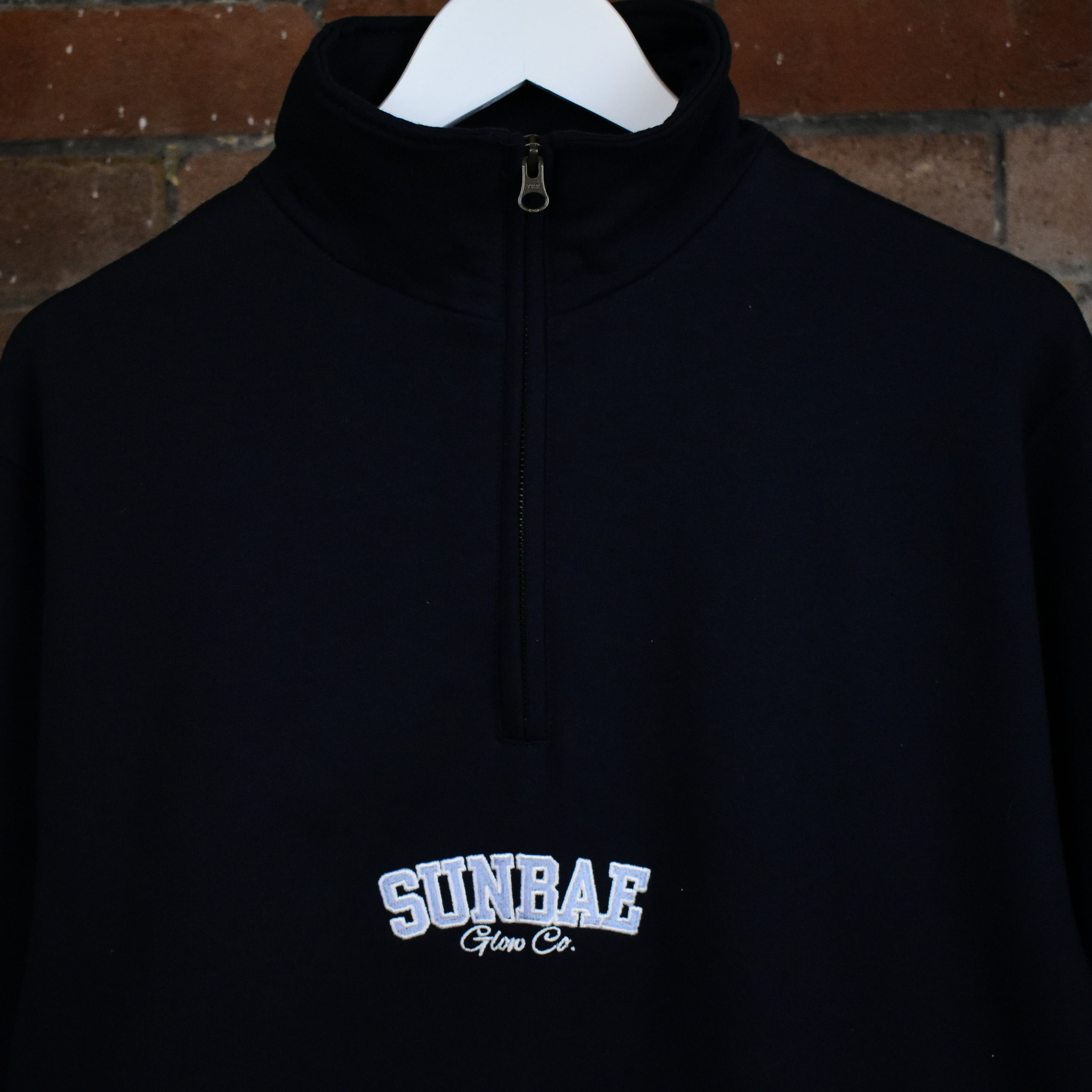 Navy hoodie with 'SUNBAE' logo on a hanger against a brick wall.