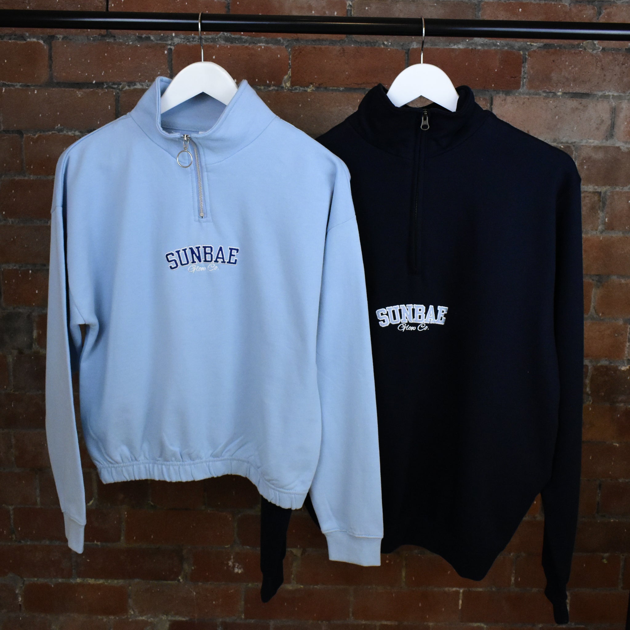 Two hoodies, one light blue and one black, hanging against a brick wall with 'SUNBAE' branding.