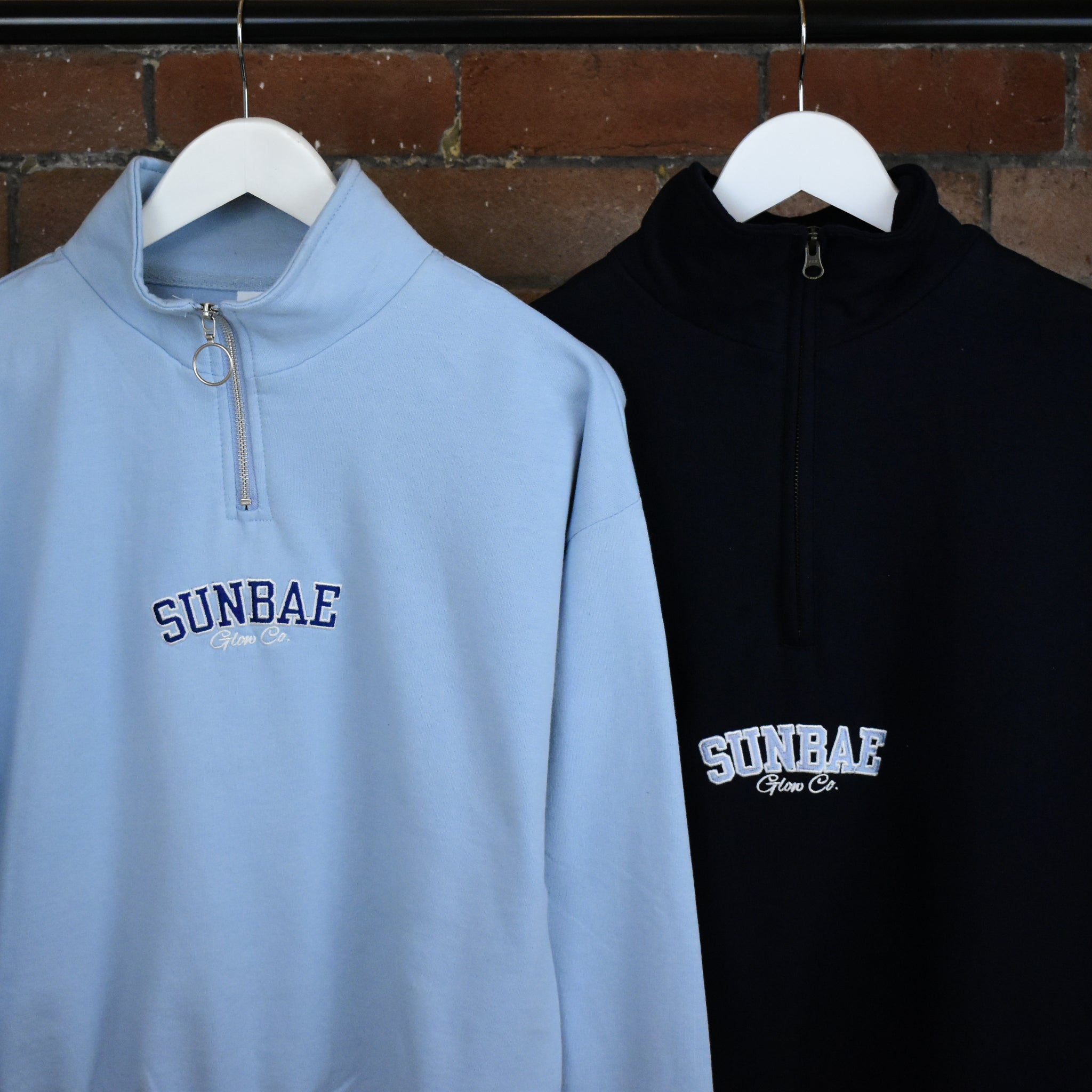 Light blue and black quarter-zip sweatshirts with 'SUNBAE' branding on hangers against a brick wall.